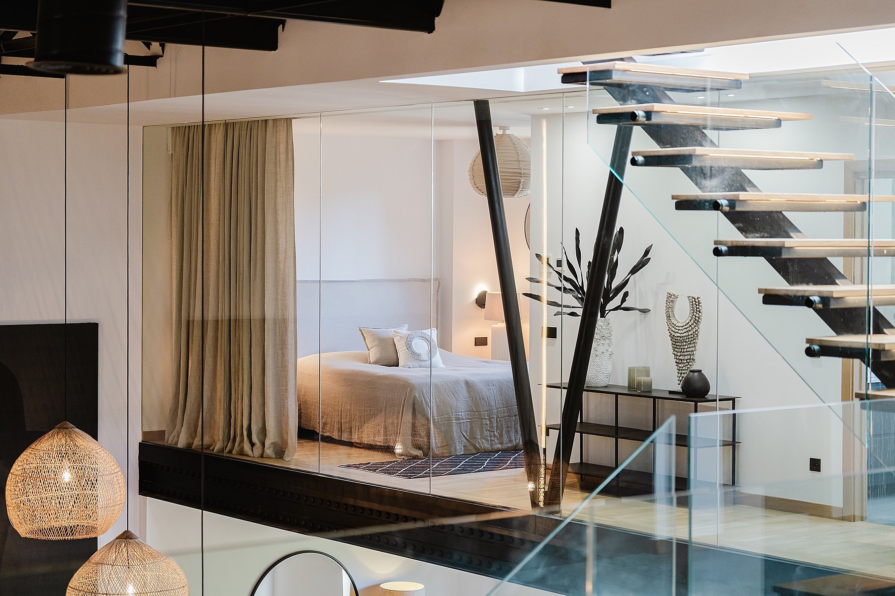 Interior view showing a master bedroom partially enclosed by glass partitions, featuring a modern floating staircase with glass railings and dark steel beams.