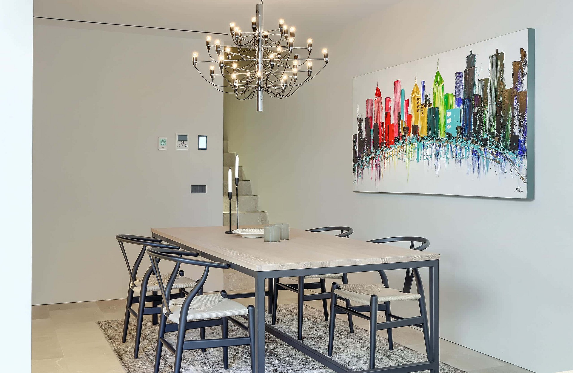 Dining room featuring a rectangular light-toned table with six black wishbone chairs on a patterned rug under a modern metal chandelier.