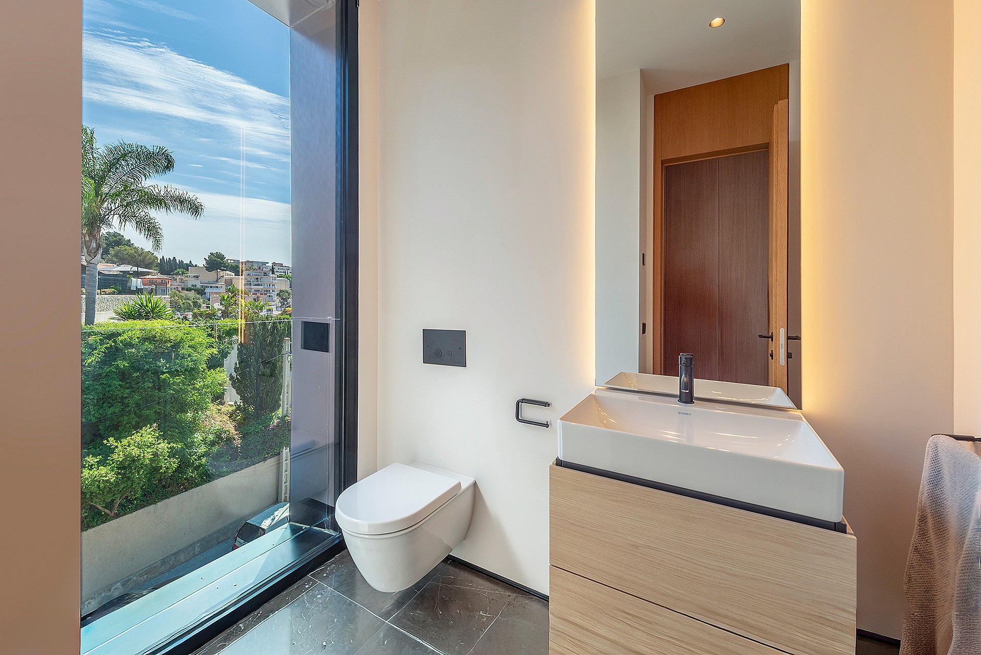 A modern guest bathroom featuring a wall-mounted toilet, wooden vanity with a white sink, backlit mirror, and floor-to-ceiling glass panel looking out to a garden.