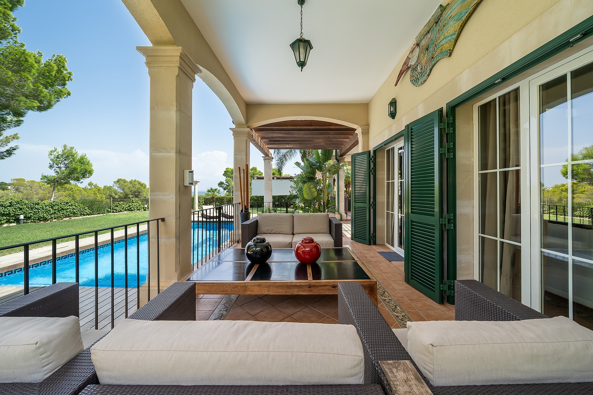 A wide covered terrace with dark rattan furniture and green louvered shutters next to a swimming pool on a sunny day.