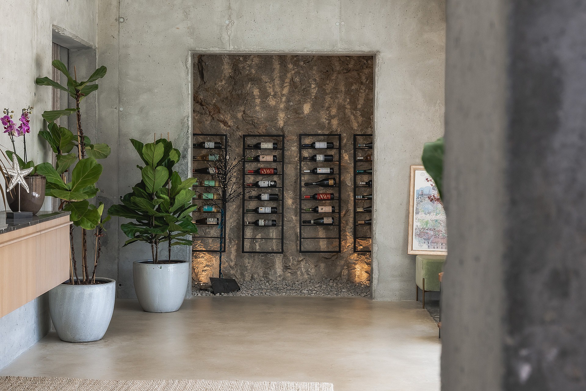 Minimalist wine storage area featuring black metal racks against an exposed rock wall and raw concrete finishes.