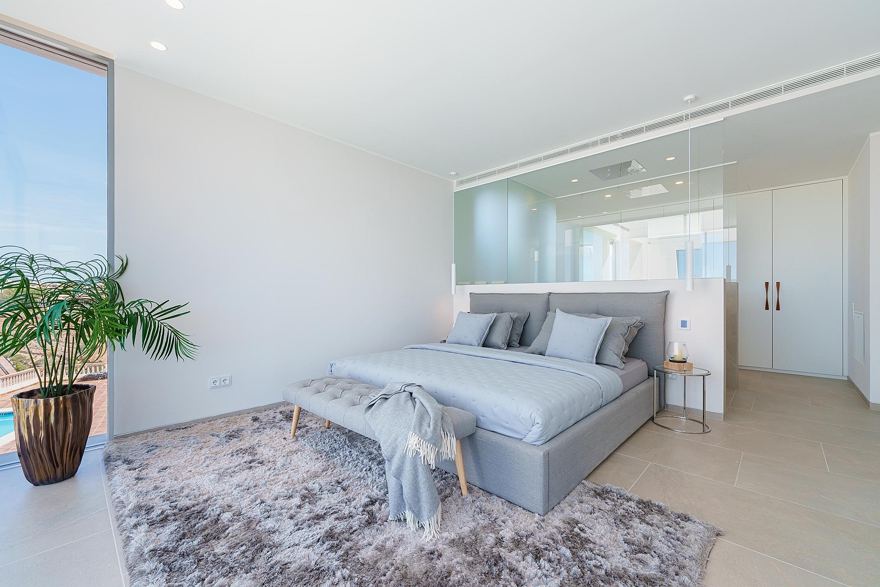 Modern bedroom with grey upholstered bed, glass-paneled en-suite bathroom, and large window overlooking a terrace.