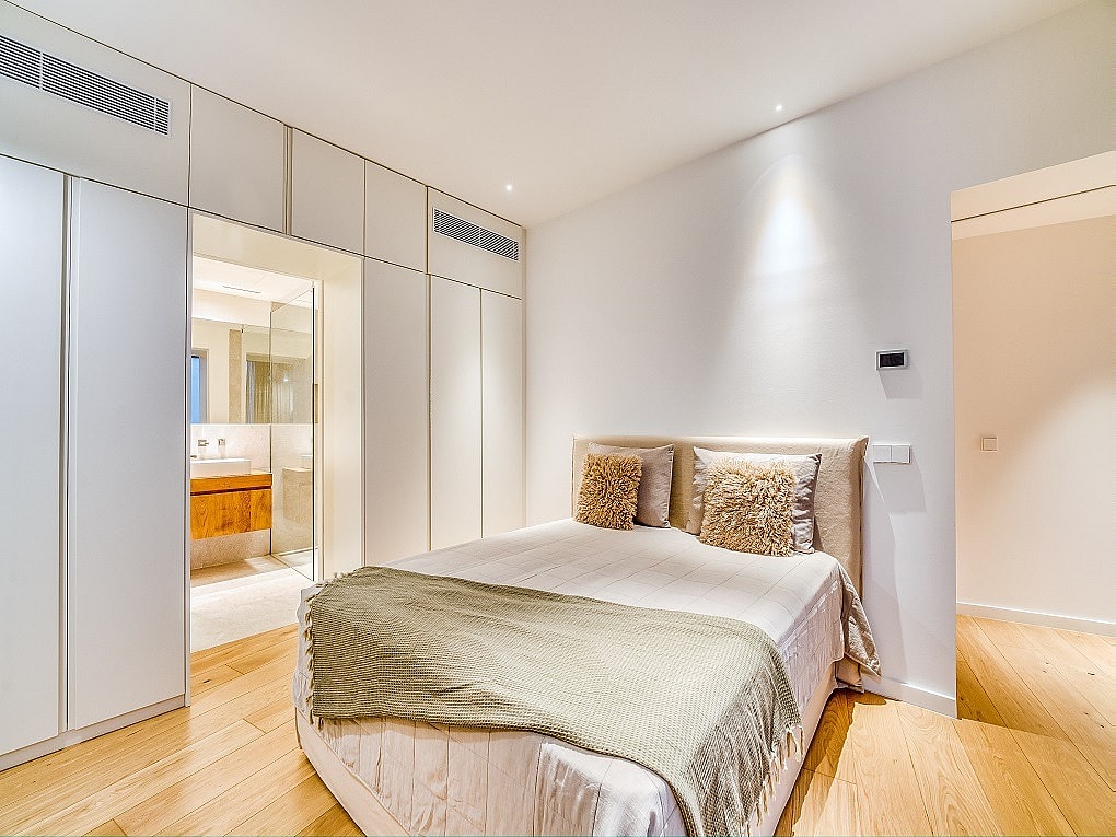 A minimalist bedroom featuring floor-to-ceiling white wardrobes, light oak wood flooring, and a bed with textured cushions.