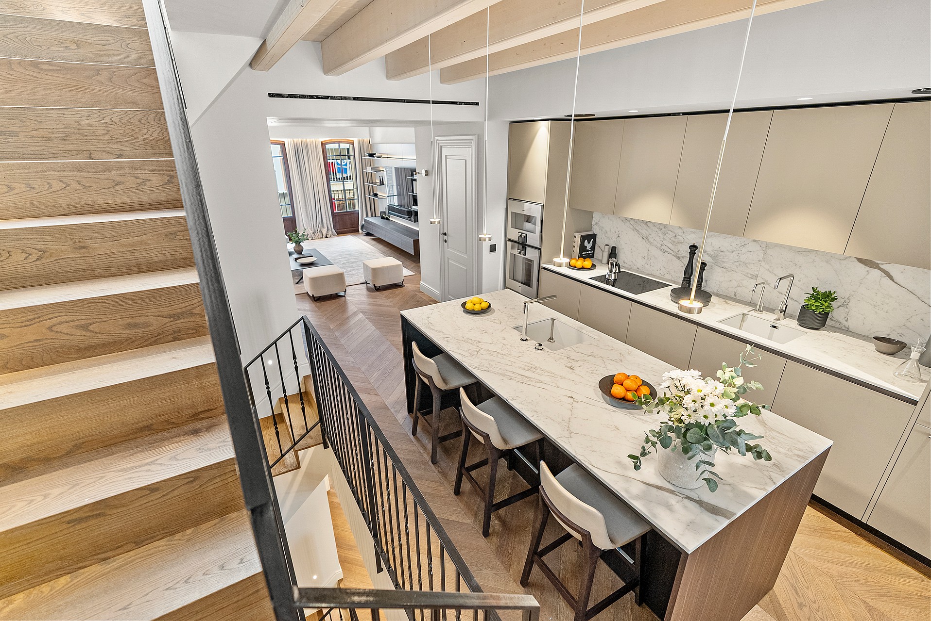 High-angle view of a modern kitchen featuring a large marble island, chevron oak floors, and stairs with metal railings.