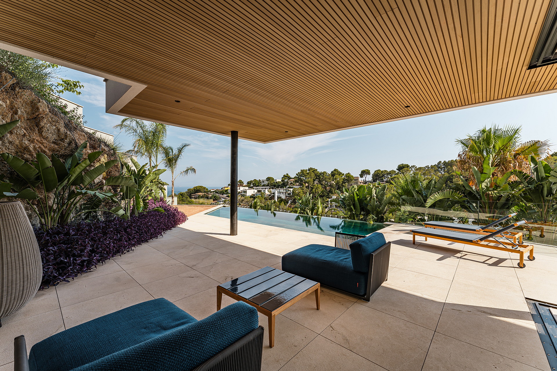 A covered terrace with a wood-slatted ceiling, limestone flooring, and blue outdoor lounge furniture overlooking a swimming pool.