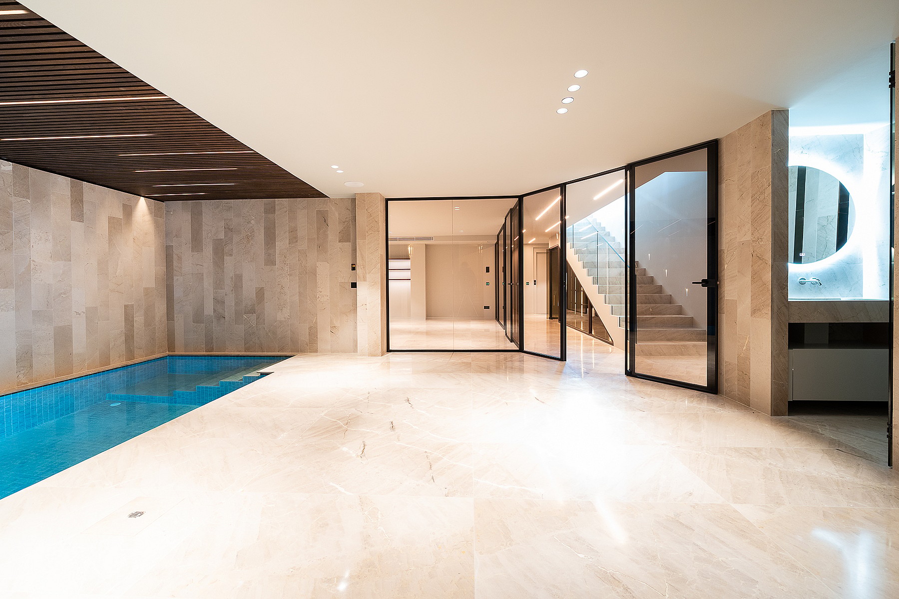 An indoor swimming pool with blue water and beige travertine flooring, adjacent to a glass-enclosed staircase.