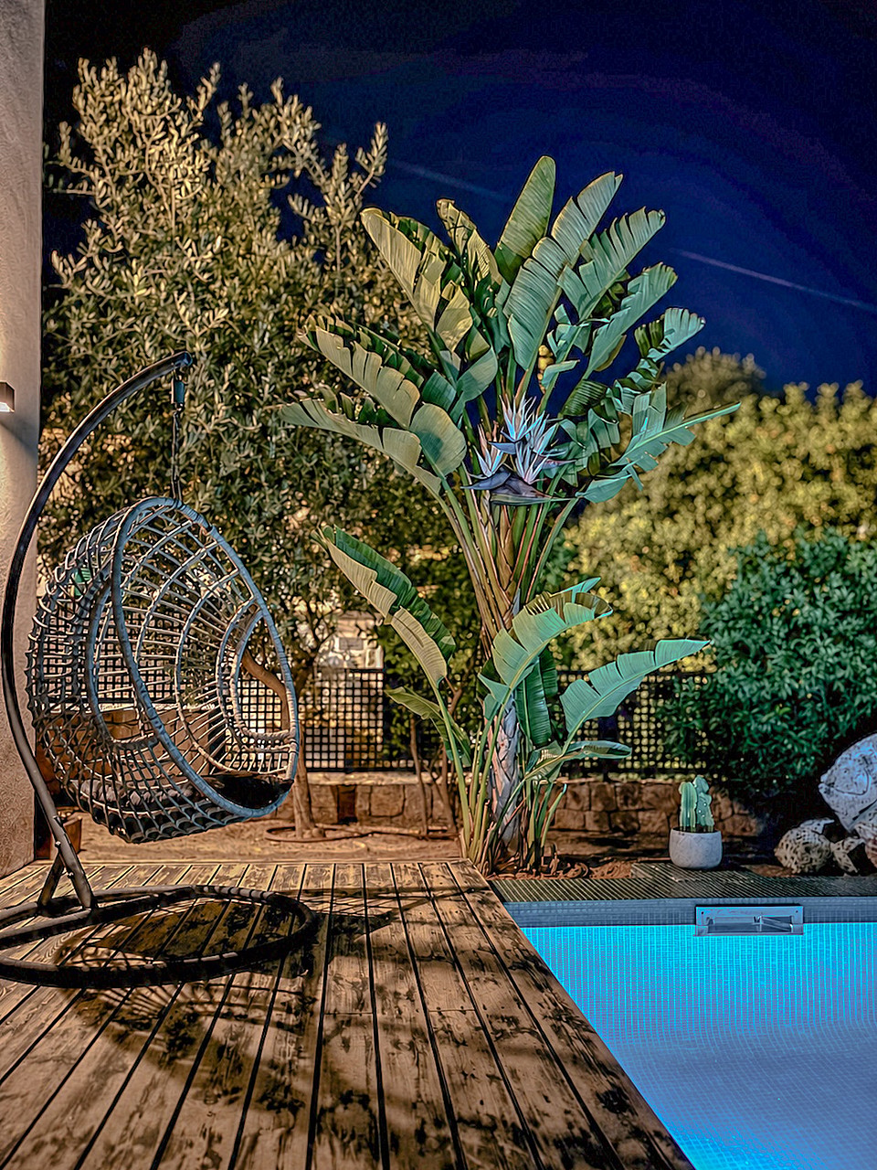 A poolside wooden deck at night with an illuminated swimming pool, a hanging wicker chair, and lush tropical foliage.