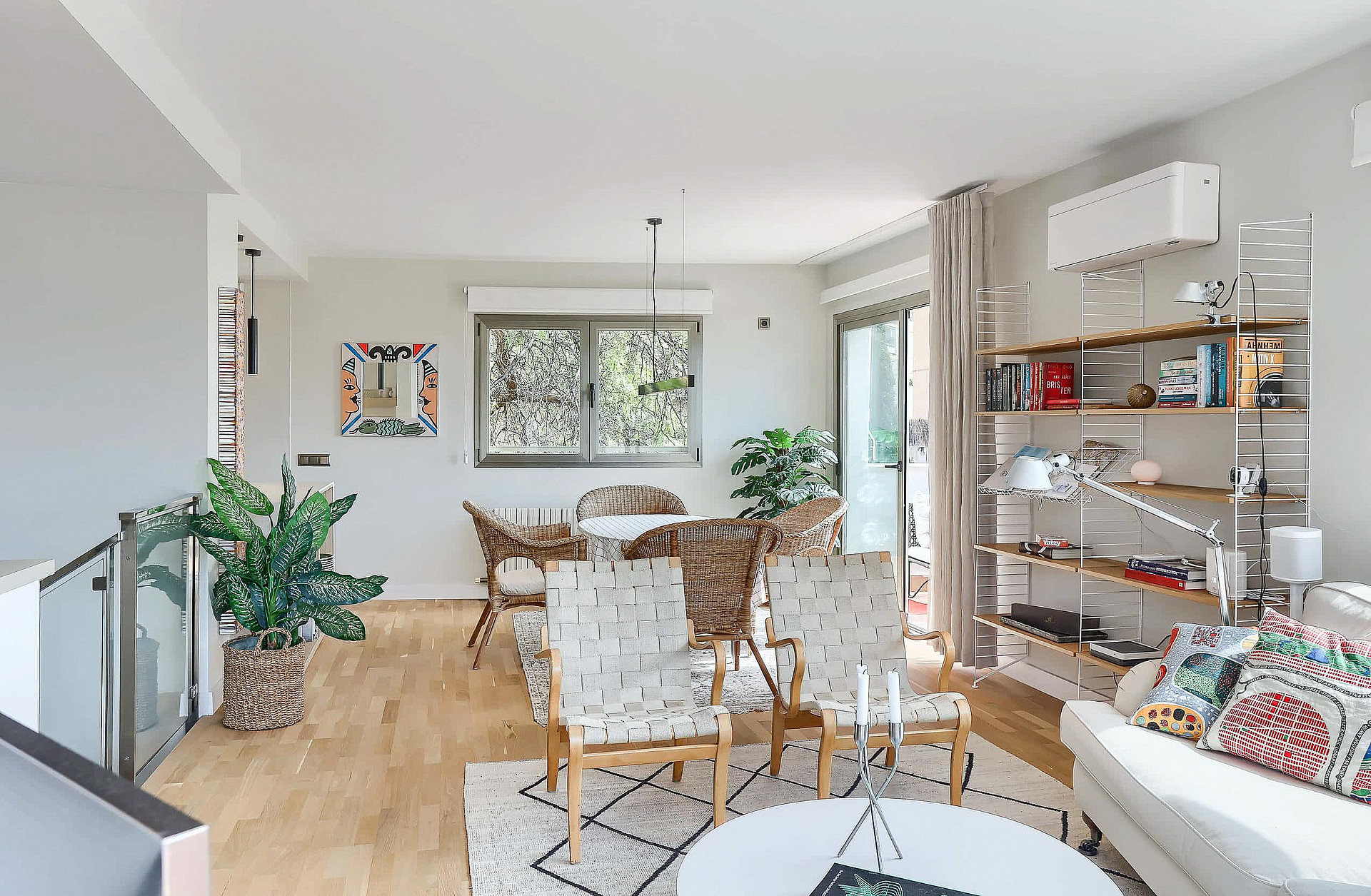 An open living room and dining space with light wood flooring, contemporary furniture, a patterned rug, and potted plants.