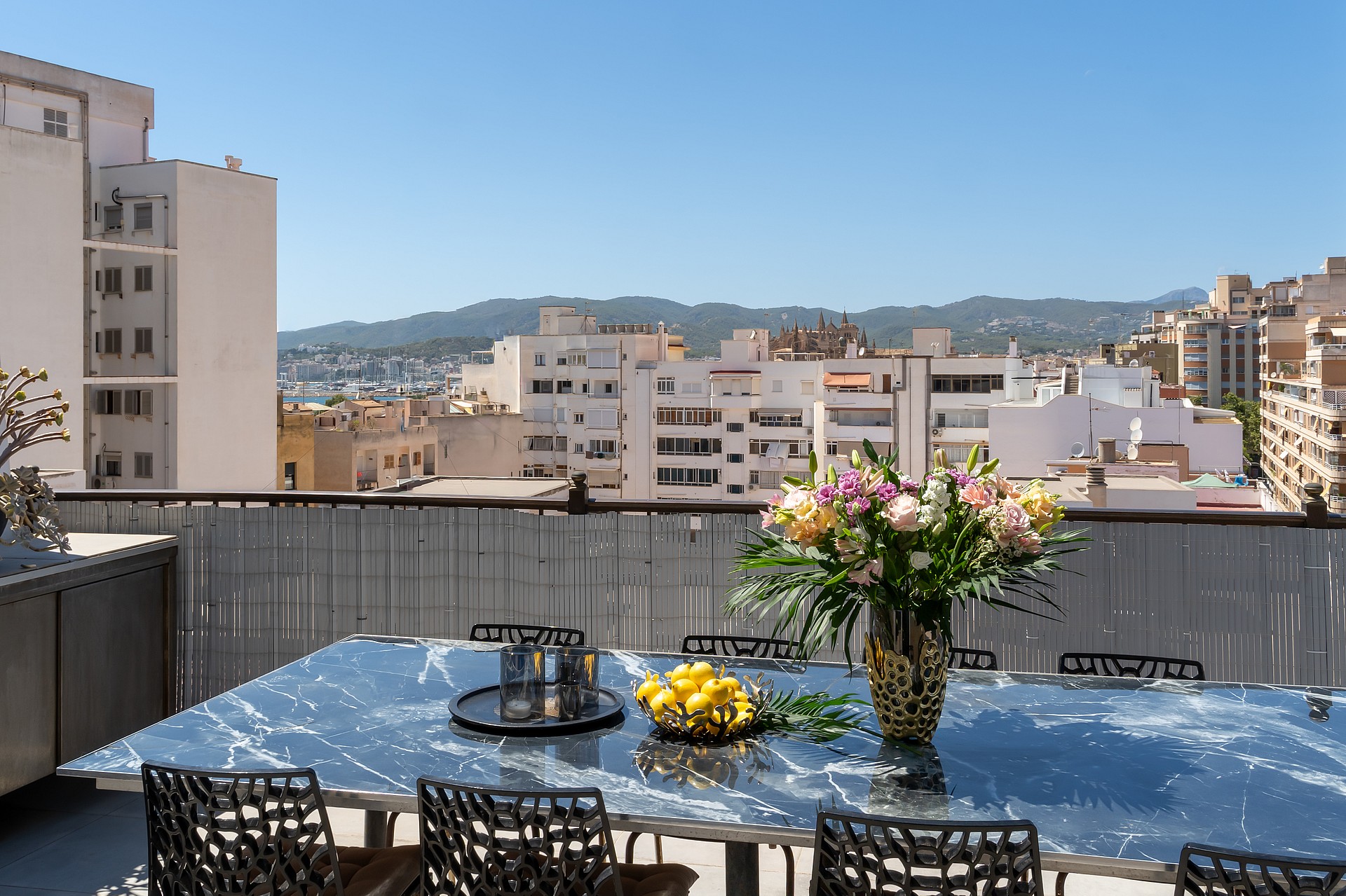 A rooftop terrace with a reflective glass dining table, black wicker chairs, and a flower vase, overlooking Old Town buildings.