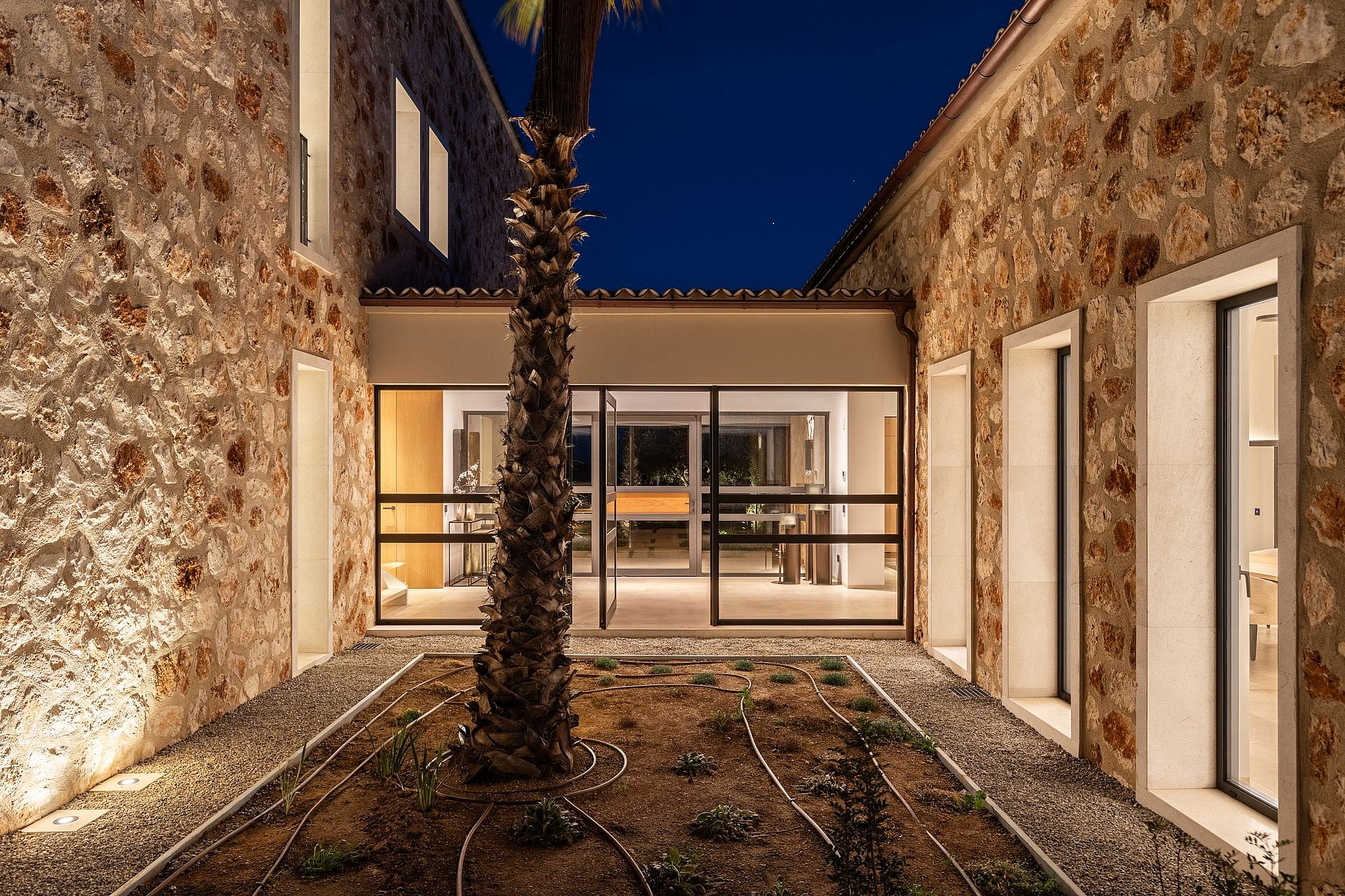 A central courtyard in a stone-clad finca at dusk, featuring a palm tree in a gravel bed and illuminated glass doors.