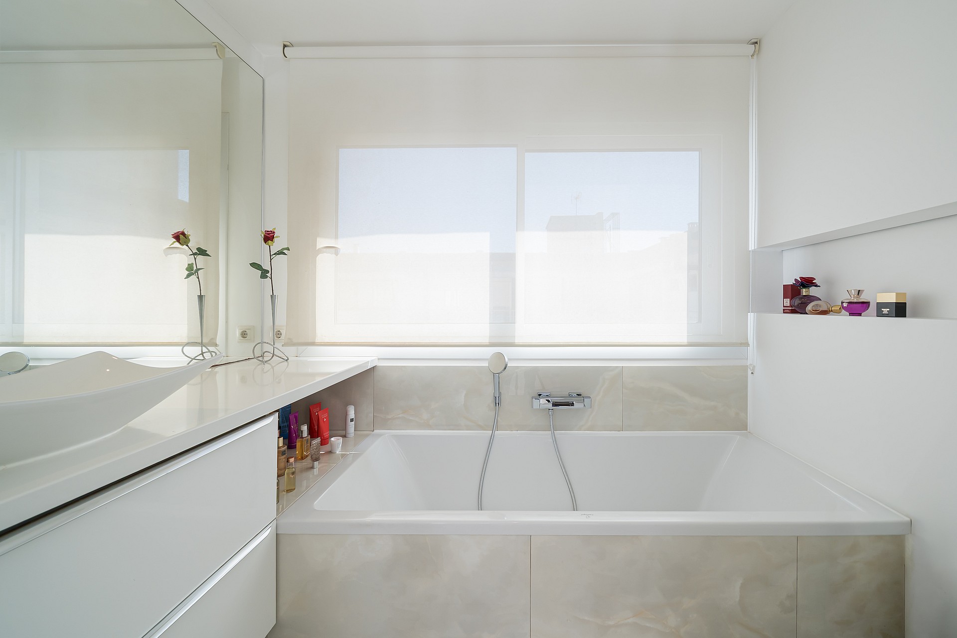 A white bathroom featuring a deep rectangular soaking tub, a wide vanity, and a large window with a roller blind.