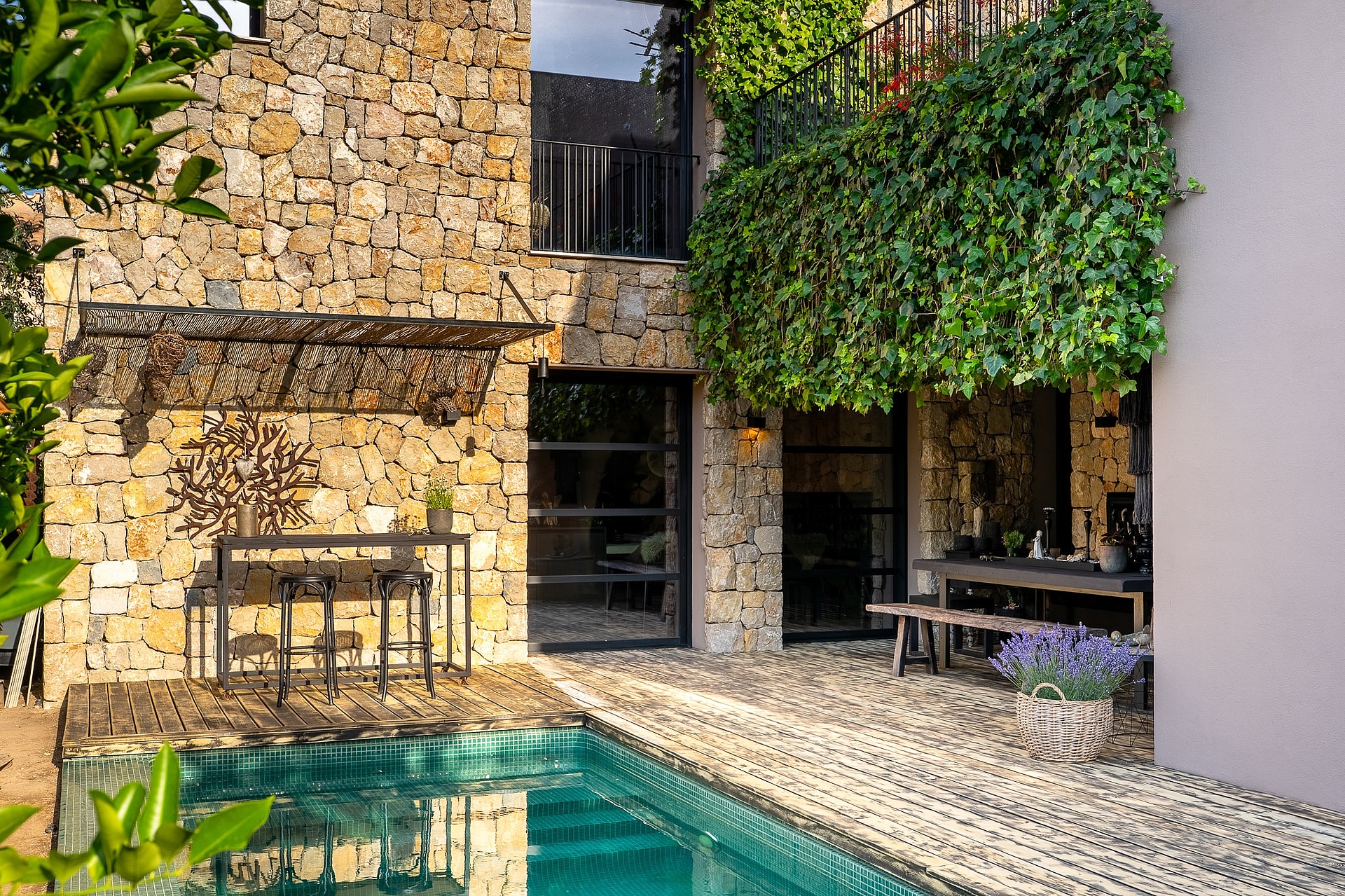 A stone villa facade with a swimming pool, outdoor dining table, and climbing greenery on the terrace wall.