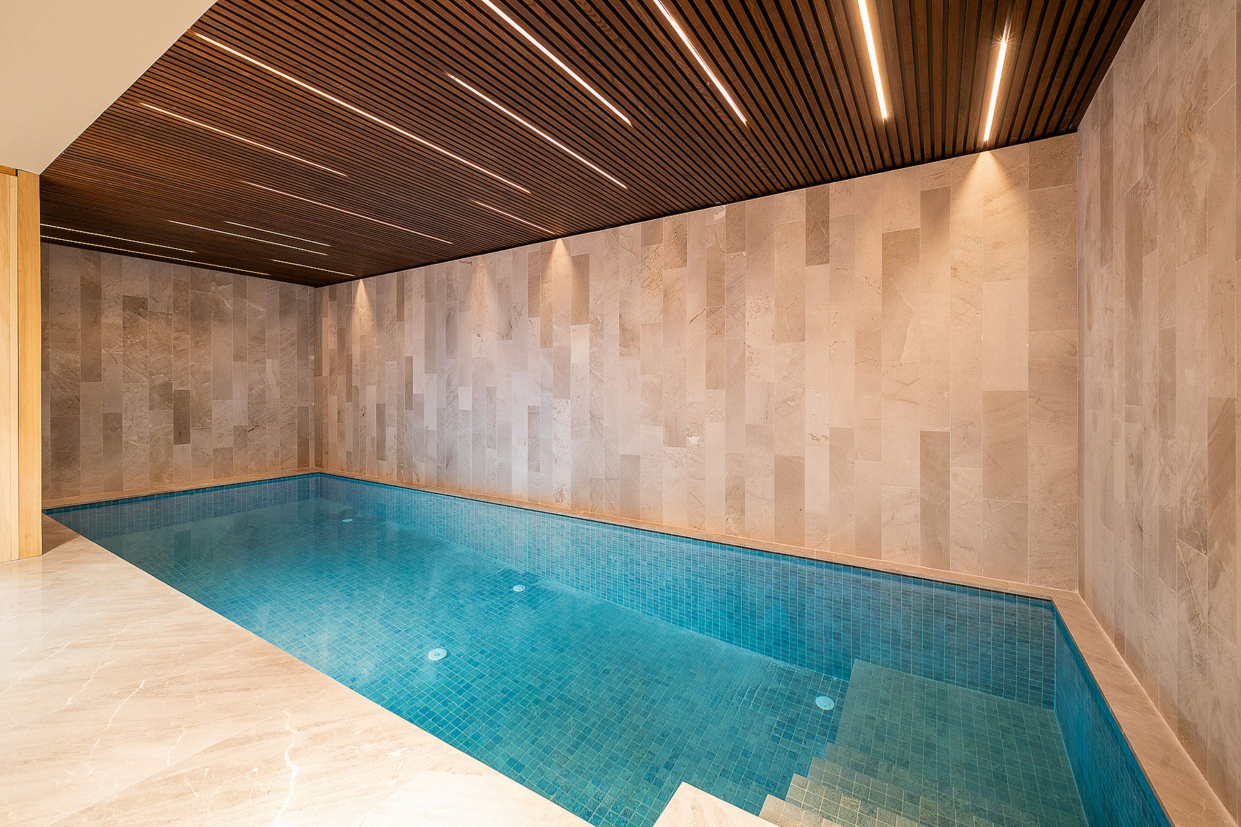 Indoor pool with turquoise water, neutral stone wall cladding, and dark wood slat ceiling with recessed linear lighting.