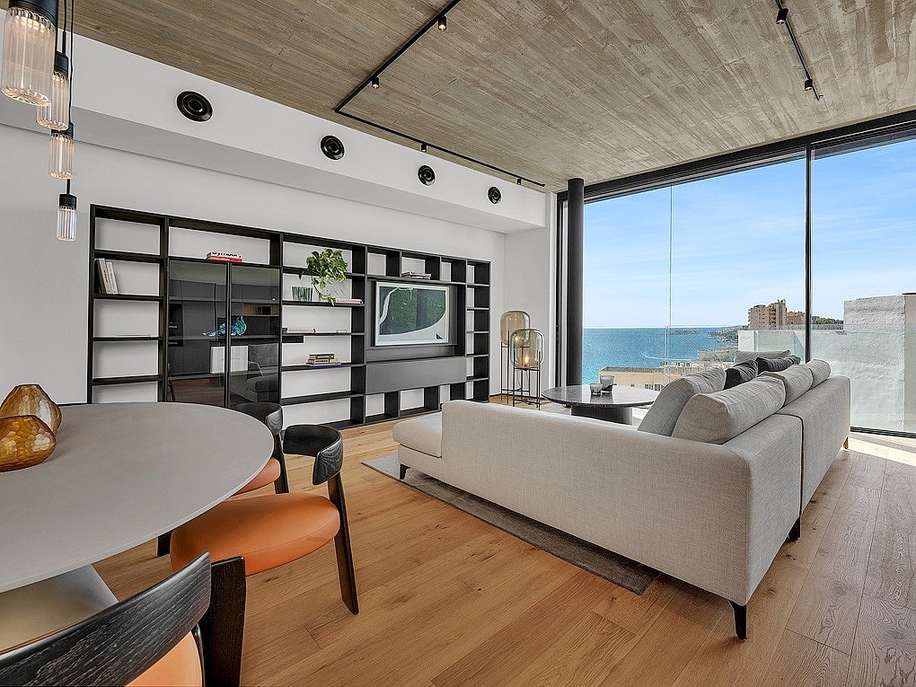 Modern living room featuring a concrete ceiling, oak flooring, large floor-to-ceiling windows overlooking the sea, and a minimalist black shelving unit.
