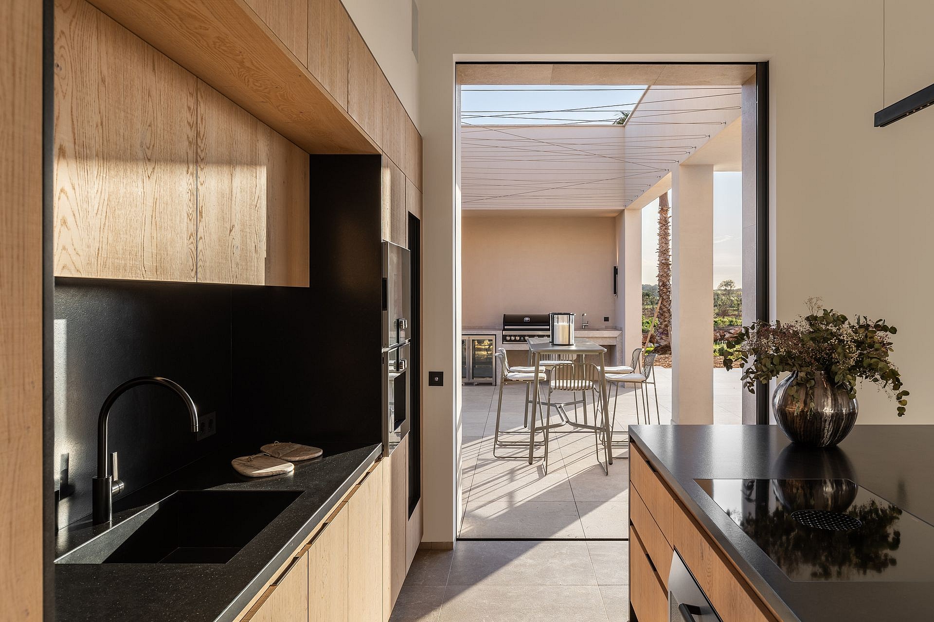 Modern kitchen with natural wood cabinetry and black stone countertops leading through a large opening to an outdoor patio.