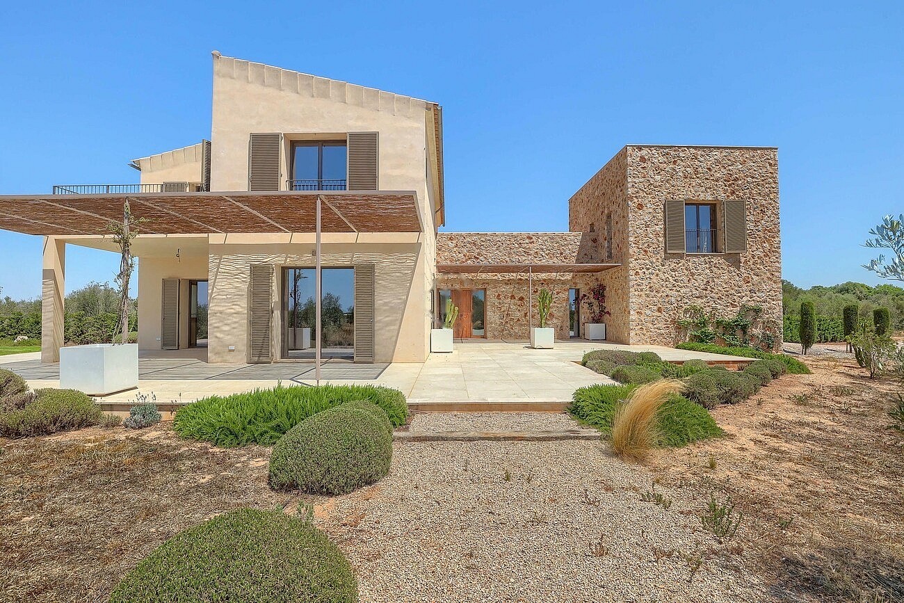 Exterior view of a Mediterranean stone finca featuring a wooden pergola, gravel path, and landscaped garden under a clear sky.