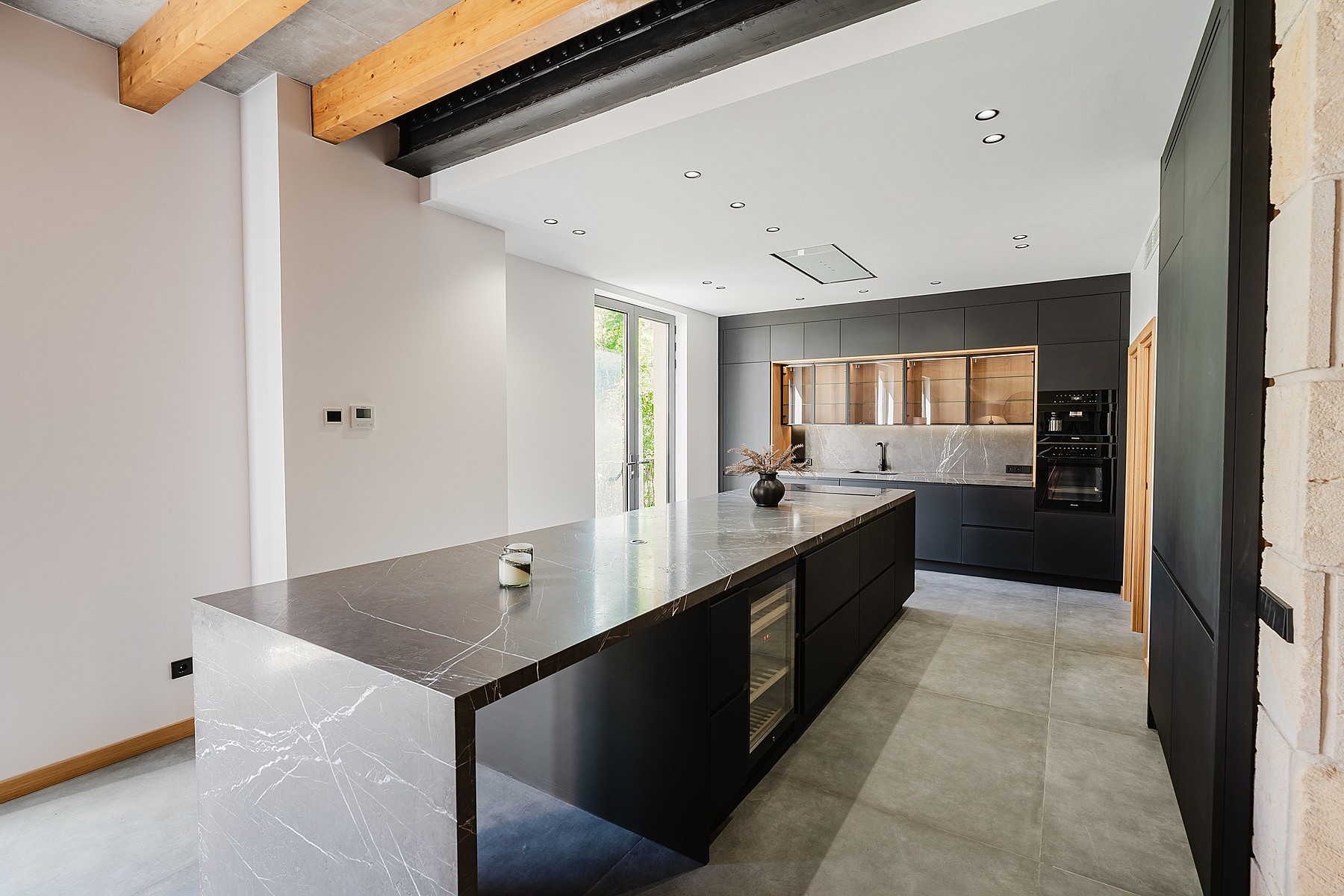 Modern open-plan kitchen in an apartment featuring a large dark stone island with waterfall edges, matte black cabinetry, and exposed wooden ceiling beams.