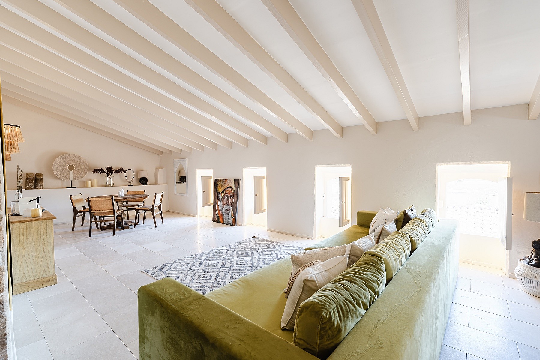Spacious living room featuring a long olive green sofa, white stone flooring, and exposed wooden beams on a slanted ceiling.