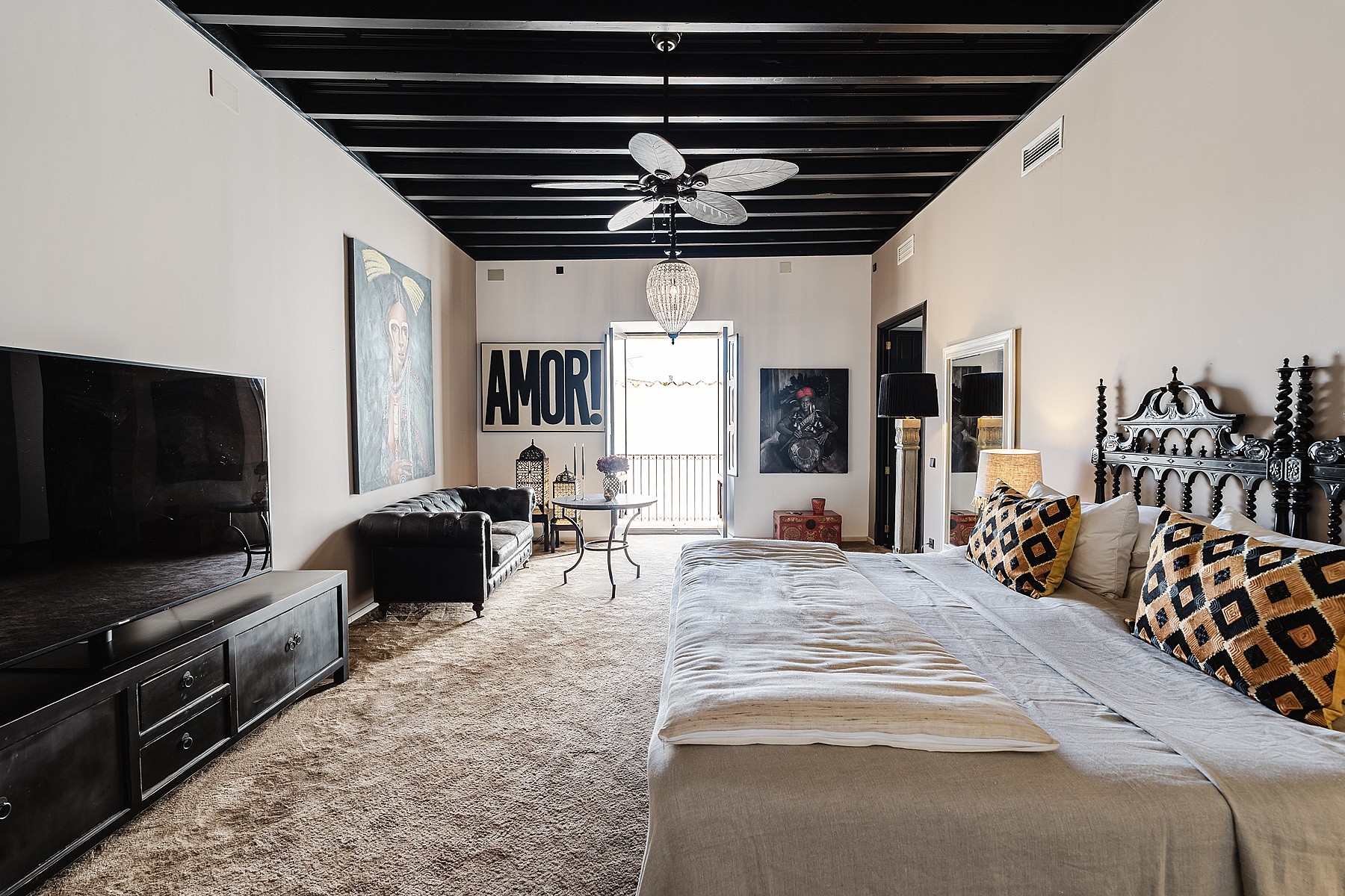 A spacious master bedroom with a white bed, black exposed wood beams, and a small seating area leading to a balcony.