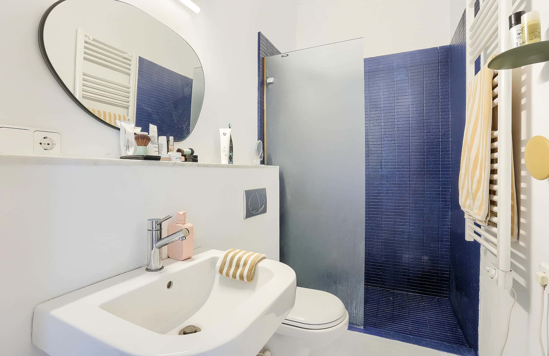 Interior view of a compact bathroom featuring a white sink, an oval wall mirror, and a walk-in shower with cobalt blue tiles.