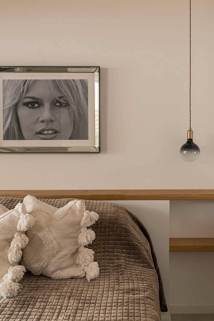 A close-up of a bedroom interior showing a textured brown bedspread, off-white decorative pillows with tassels, and a floating light-oak headboard shelf with a pendant light.