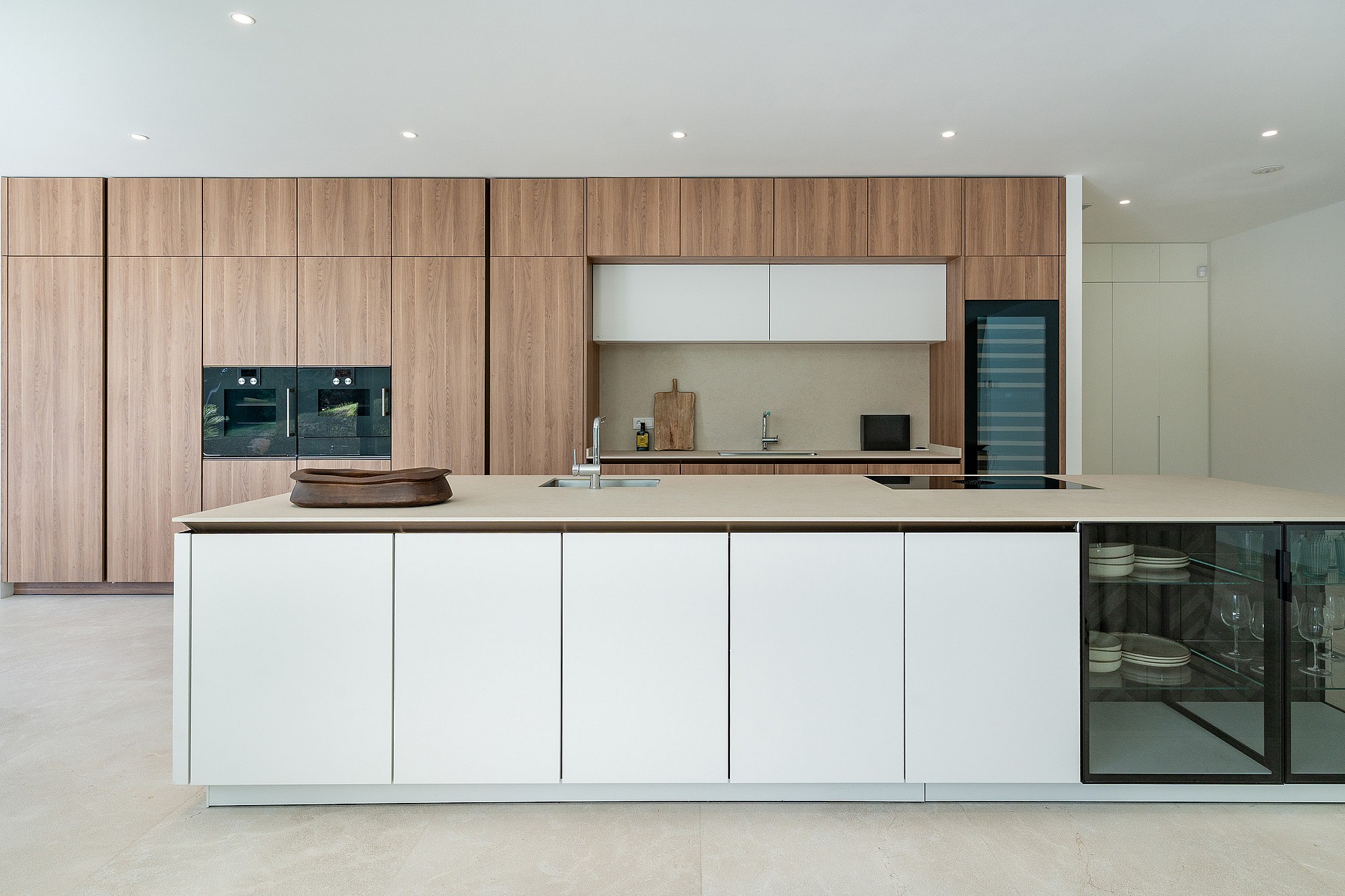White handleless Siematic kitchen island with stone worktop, integrated sink and glass-fronted crockery display, floor-to-ceiling oak-veneer cabinetry and Gaggenau ovens behind