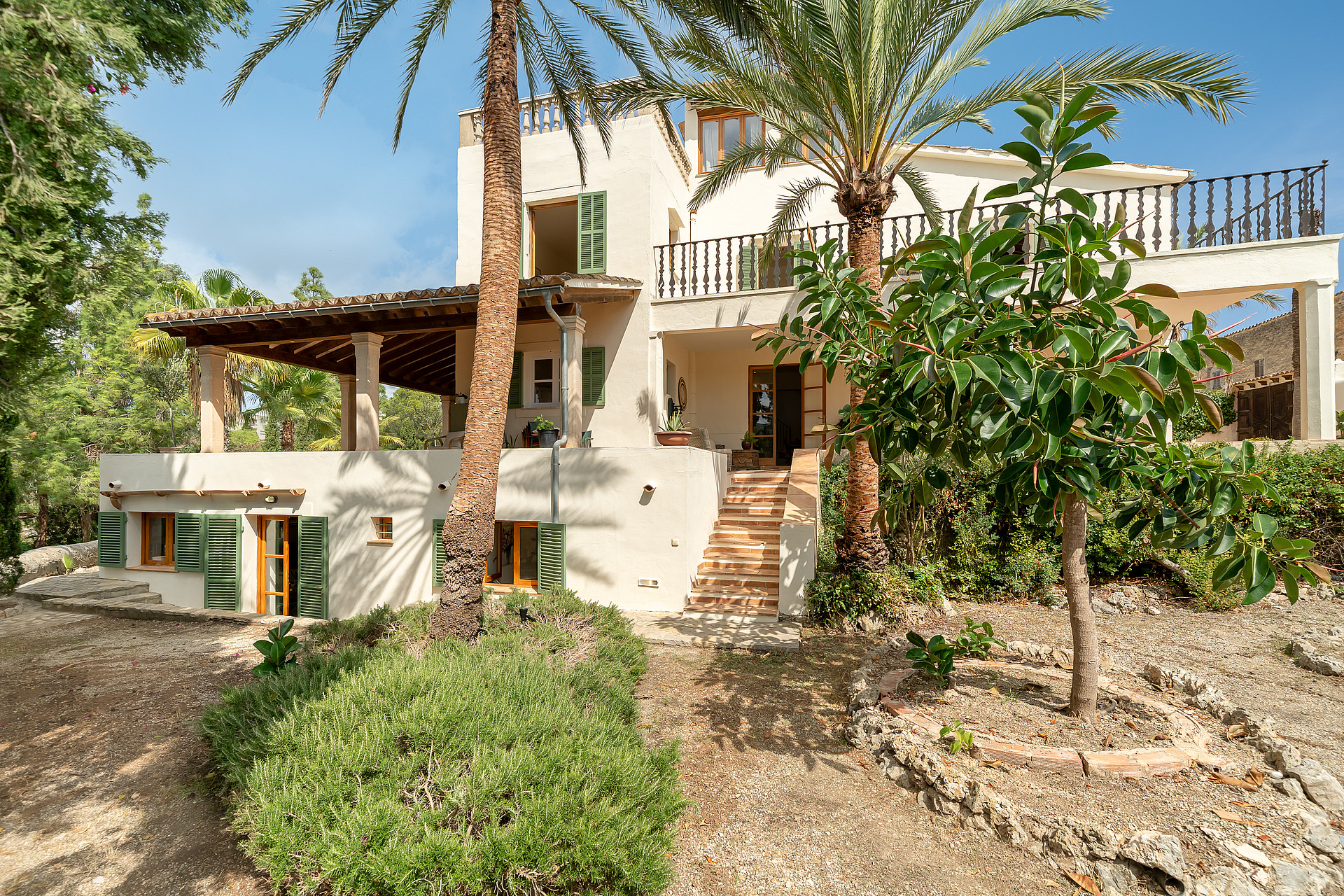 Multi-level Mediterranean villa with cream stucco walls, green shutters, and stone steps framed by palm trees.
