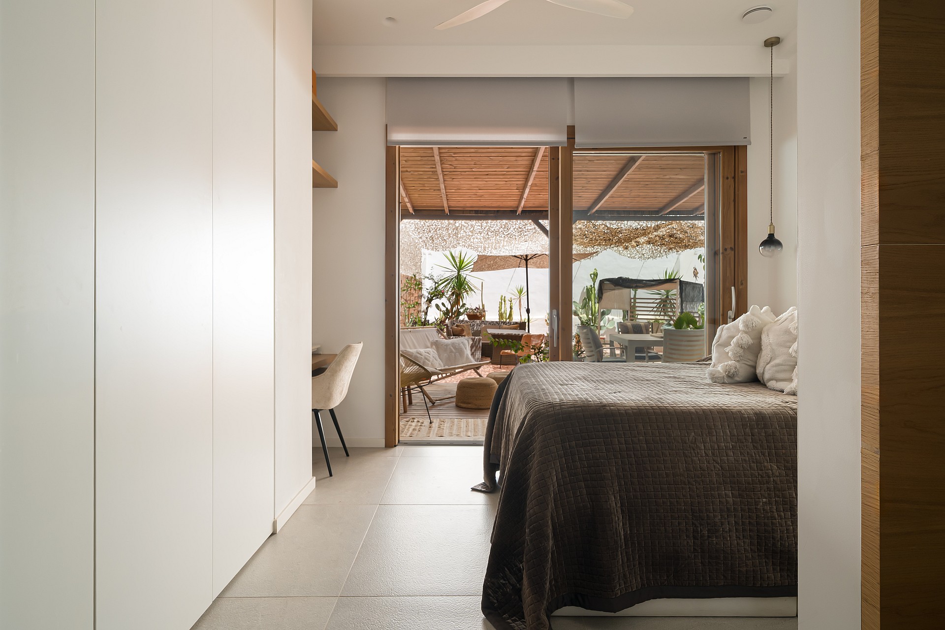 A minimalist bedroom featuring floor-to-ceiling white wardrobes, beige stone floors, and glass doors opening to a furnished terrace.