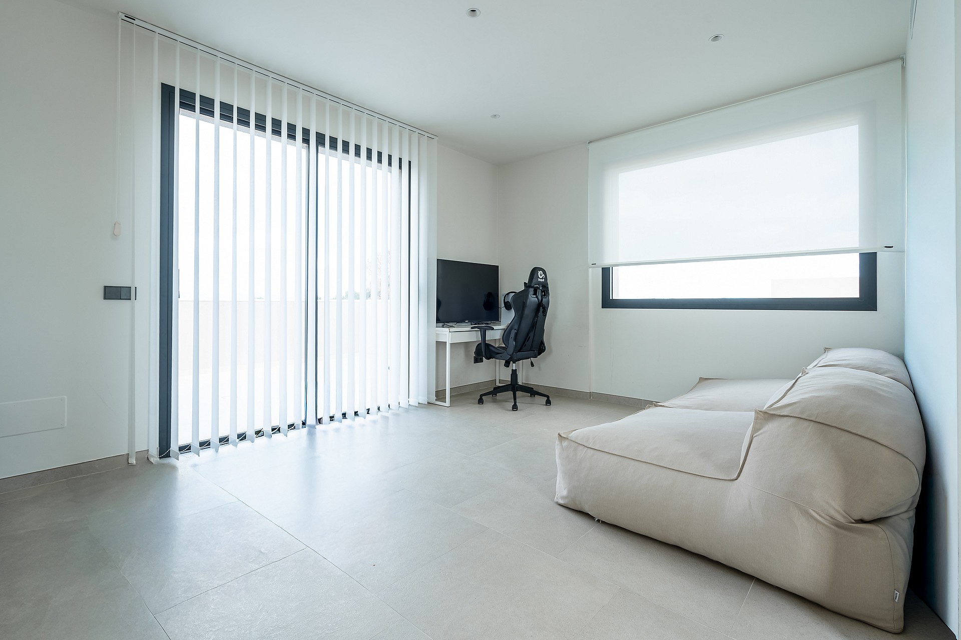 A minimalist room featuring a white desk, a black ergonomic chair, and a large beige floor cushion, with vertical blinds on a sliding glass door and polished concrete floors.