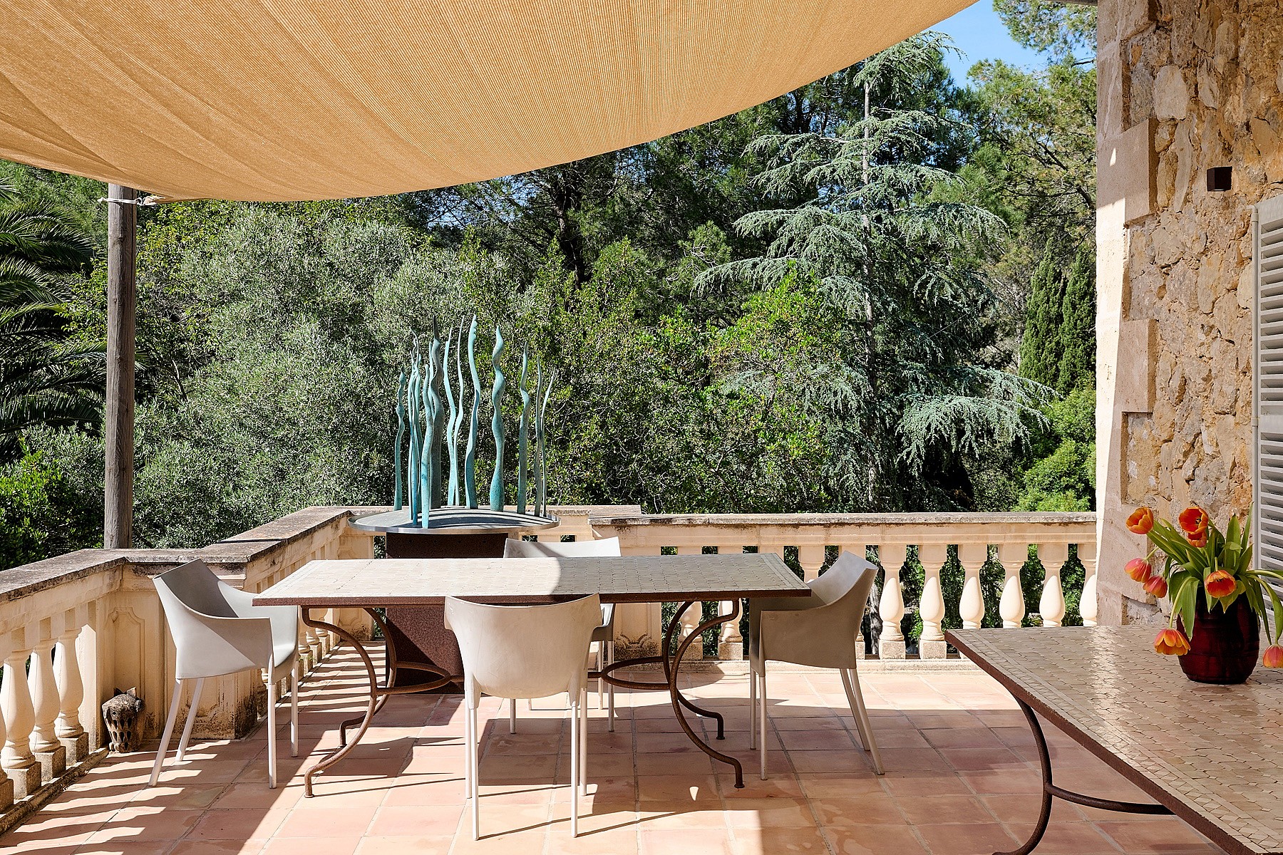 A stone terrace with a dining table and chairs, protected by a large beige sail shade, overlooking dense mountain trees.