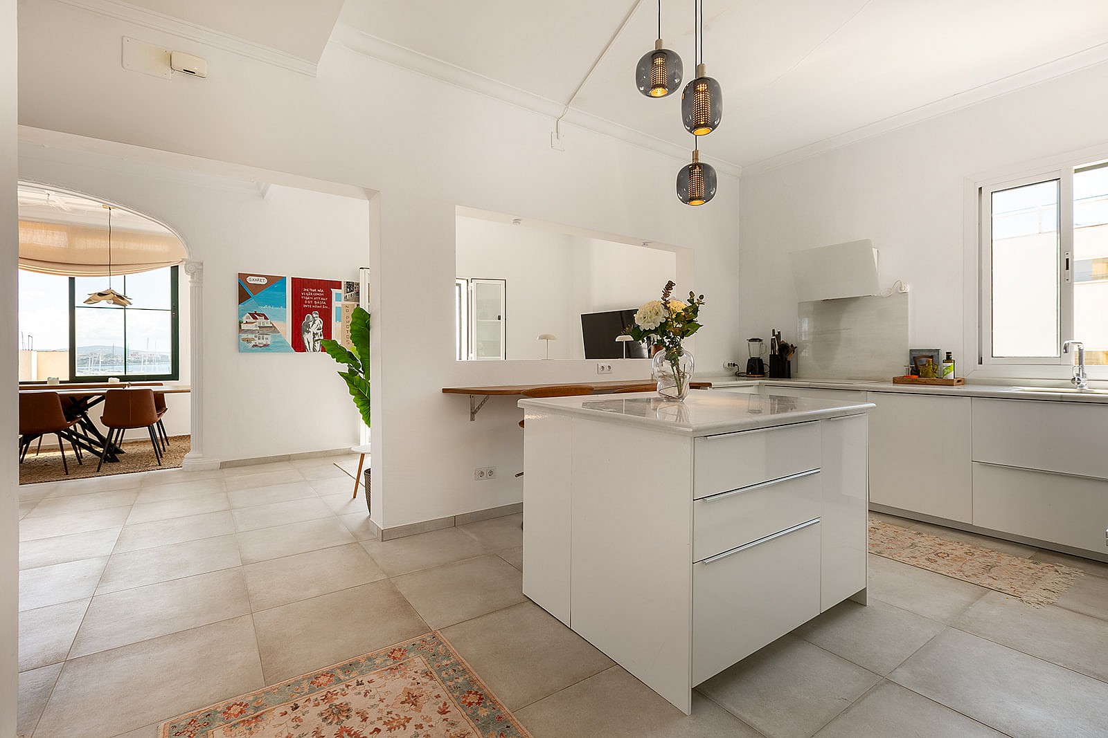 Modern kitchen with white cabinetry, a central island, and tiled floors. Pendant lights hang above the island, with an adjacent dining area visible.