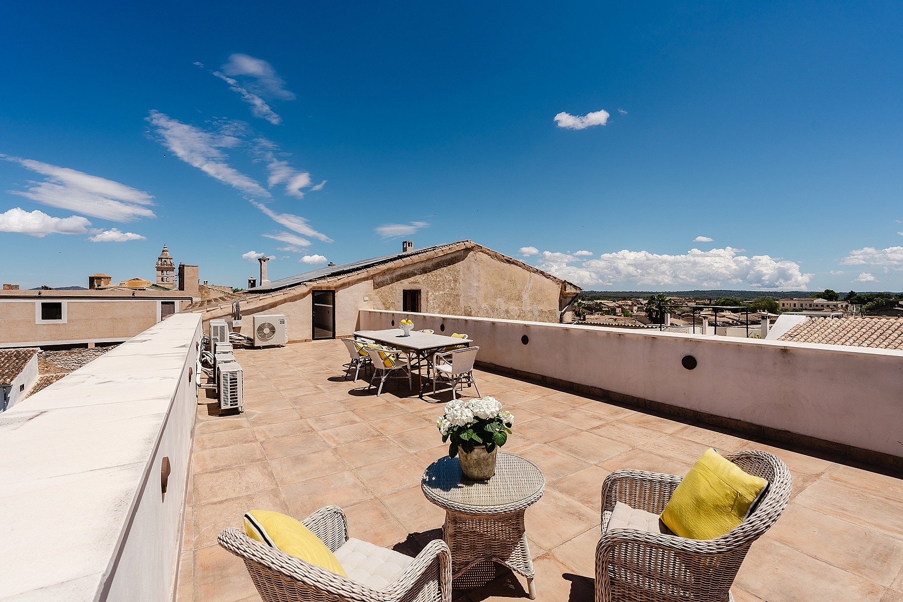 A wide rooftop terrace with stone flooring, wicker outdoor furniture, and a dining table, overlooking the village buildings.