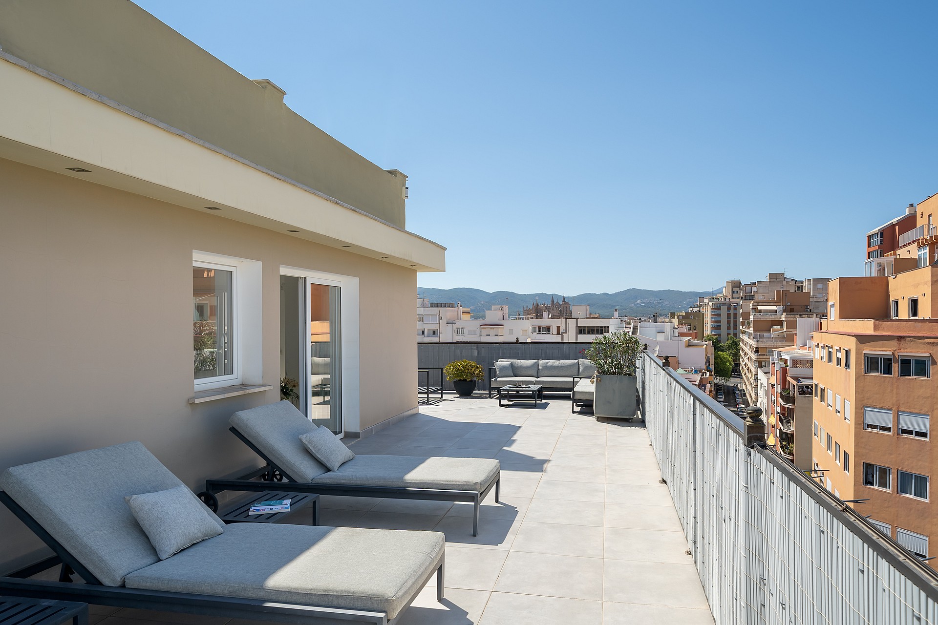 A wide rooftop terrace with light stone flooring, two grey sun loungers in the foreground, and a city skyline view.