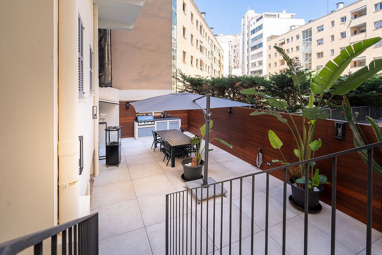 A private outdoor terrace in Palma featuring light-colored stone flooring, wooden wall panels, a dining table with chairs, and an umbrella, set against a backdrop of city buildings.