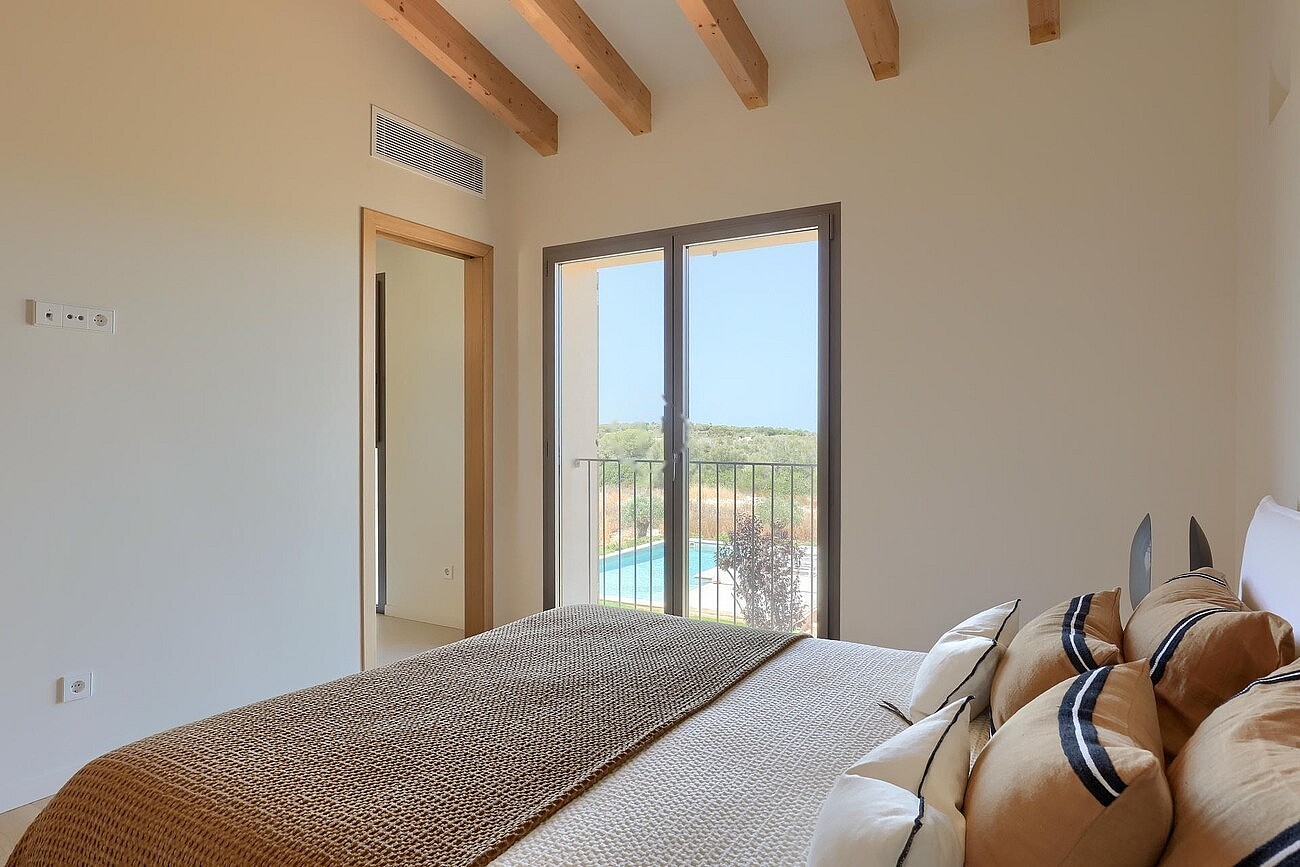 A bedroom featuring exposed timber ceiling beams, white walls, and a glass door opening to a balcony overlooking a pool.