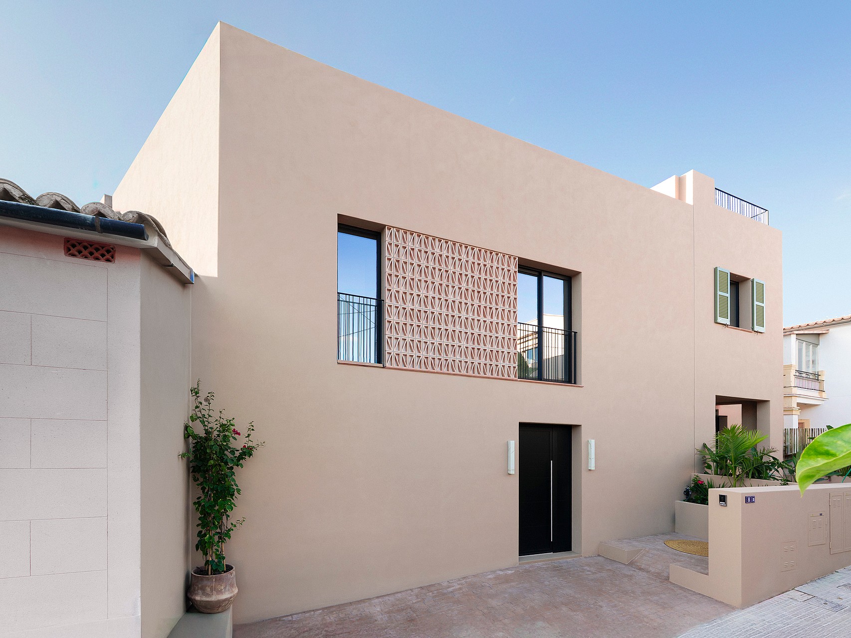 Minimalist sand-colored townhouse facade with a perforated terracotta screen, dark entrance door, and integrated planters.
