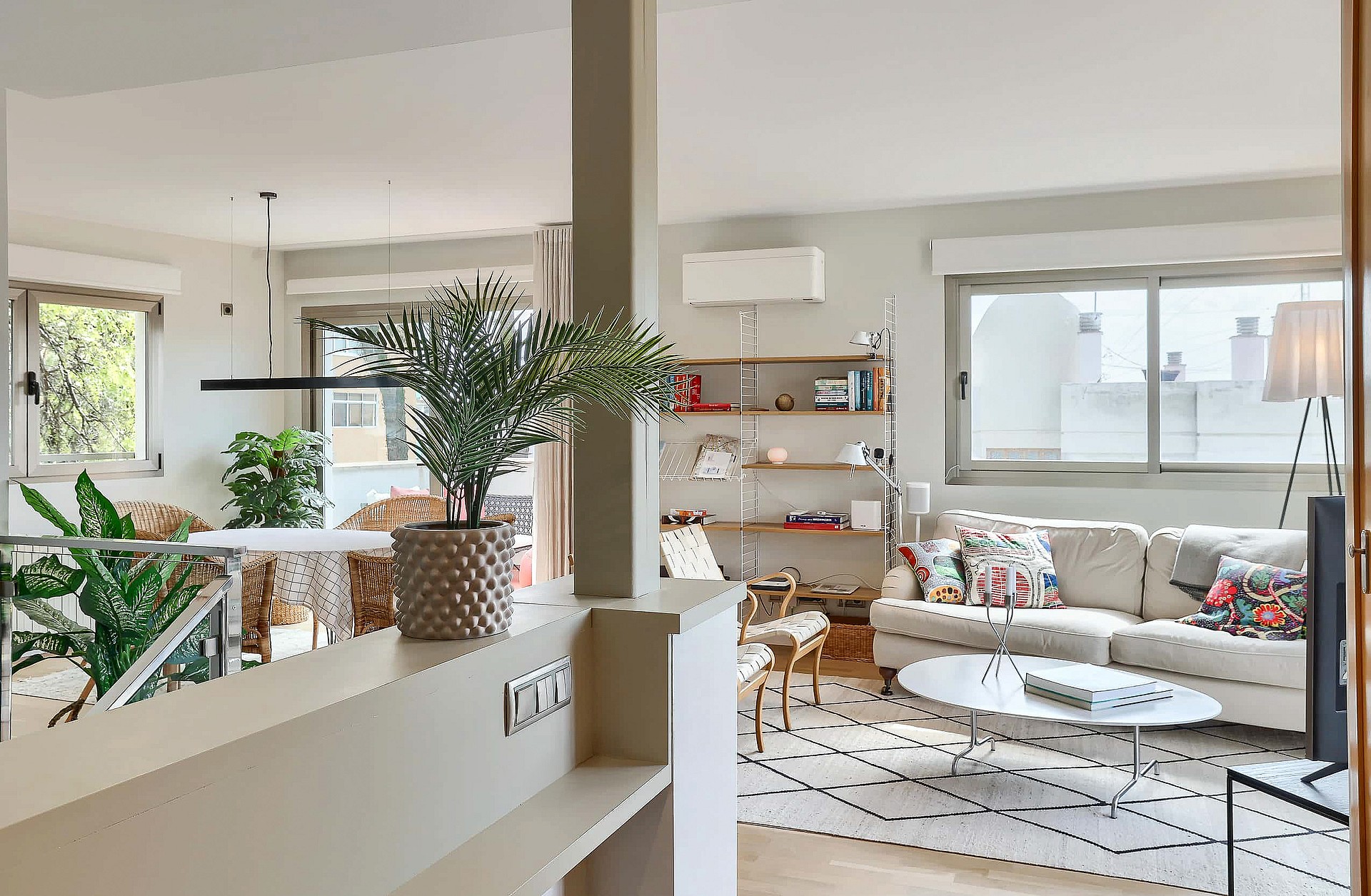 Interior view of an open-plan living room with a neutral sofa, patterned rug, open shelving, and natural light.