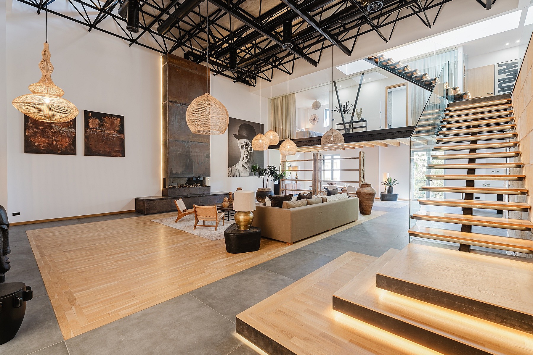 A spacious apartment interior with high ceilings, exposed metal roof trusses, a floating staircase, and neutral-toned furniture.