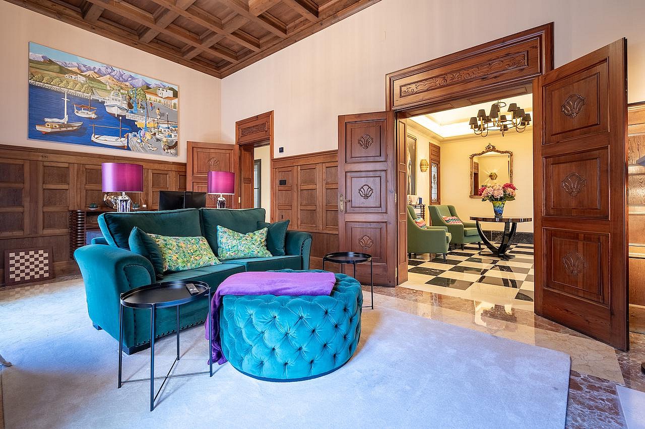 A formal living room with dark wood wall paneling, a coffered ceiling, a velvet teal sofa, and an open doorway leading to a tiled room.