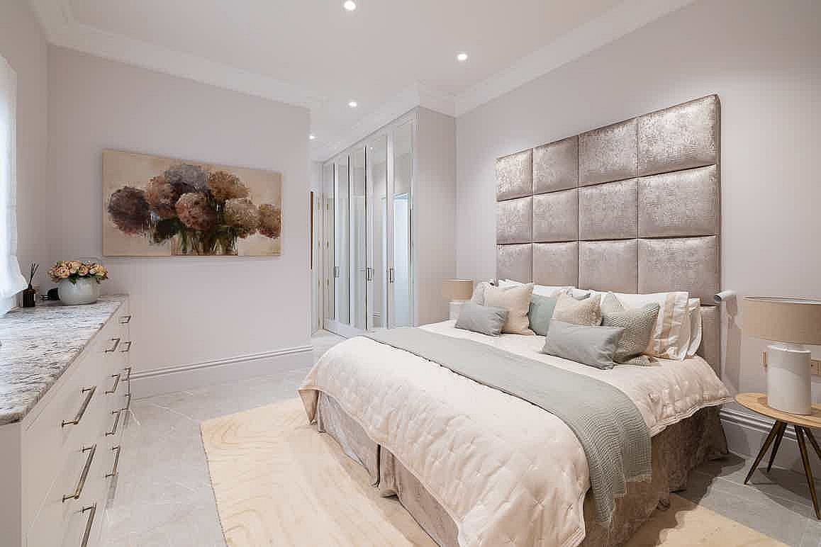 Bedroom featuring a large tufted velvet headboard, white wardrobe doors, and a floral painting on a light grey wall.