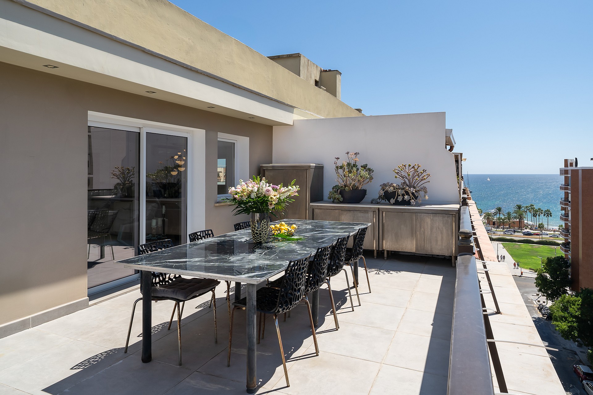 A spacious outdoor terrace with a long dining table, modern chairs, and an open view overlooking the city and the sea.