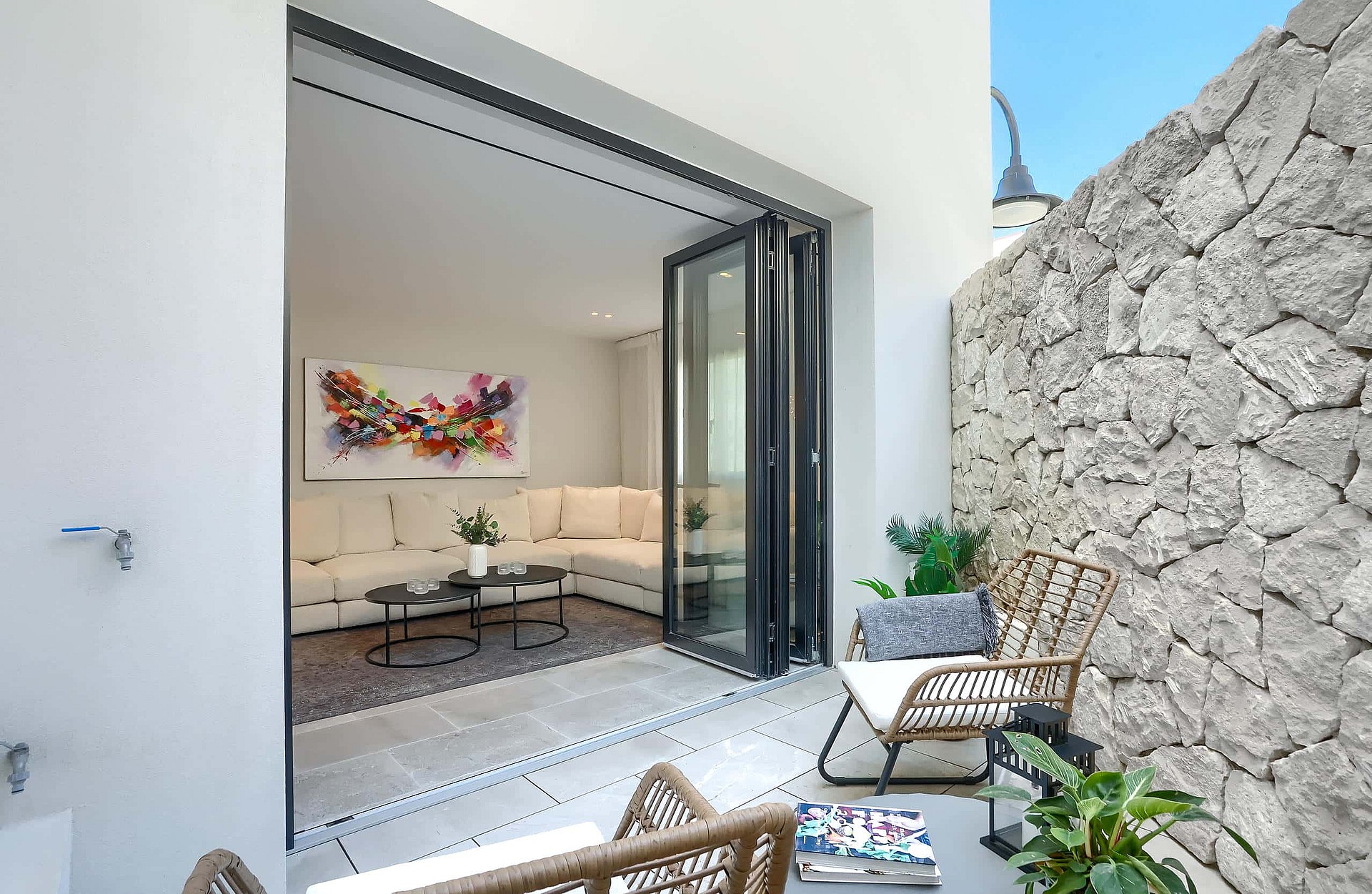A small outdoor terrace with a high natural stone wall, rattan chairs, and large folding glass doors opening into a living room.
