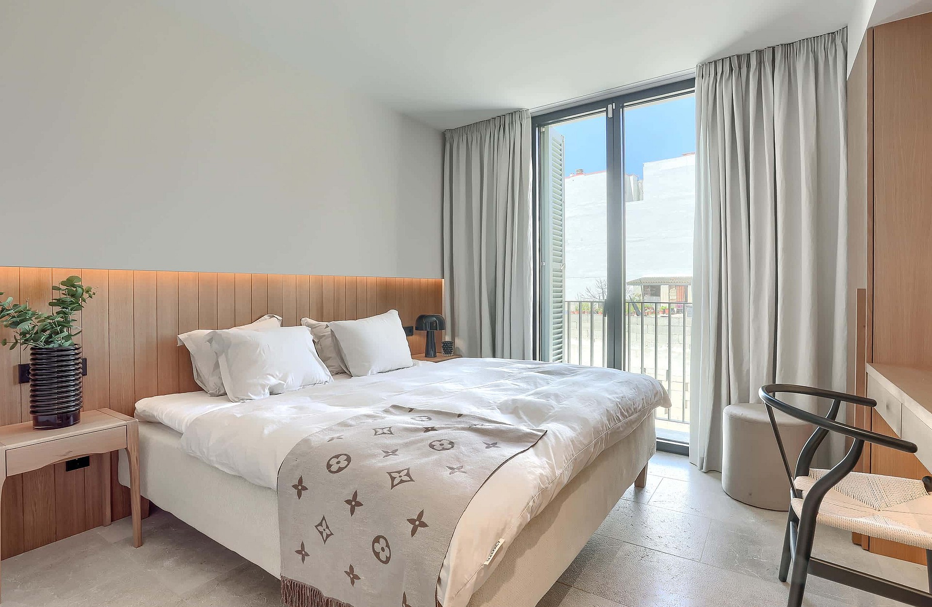 Bright bedroom with a double bed, vertical oak wood wall paneling, and floor-to-ceiling glass doors with light curtains.
