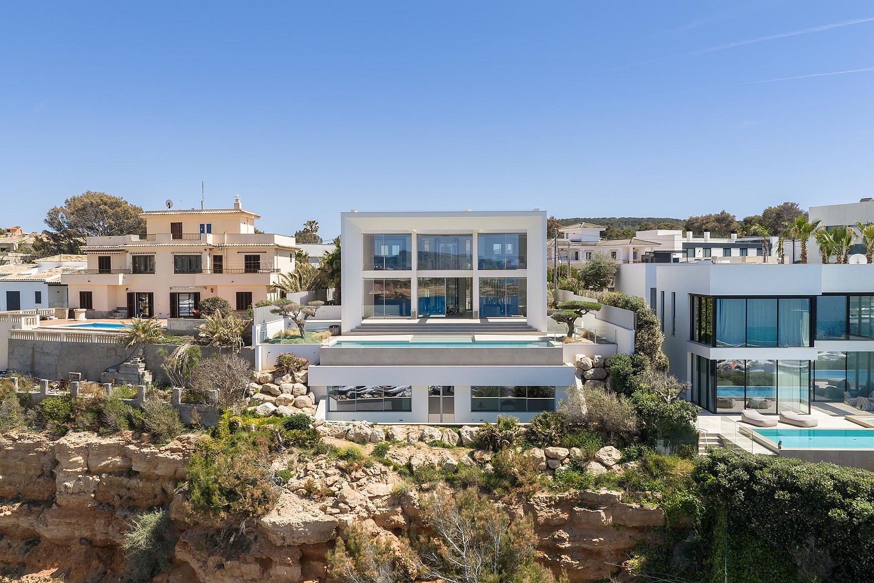 Wide exterior view of a modern white multi-level villa built into a rocky coastline with a swimming pool and glass walls.