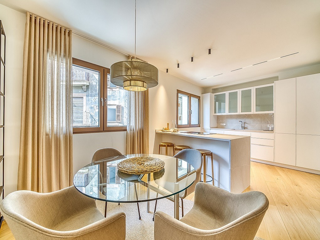 Modern open-plan kitchen with white cabinetry and dining area featuring a round glass table, beige armchairs, and floor-length curtains.