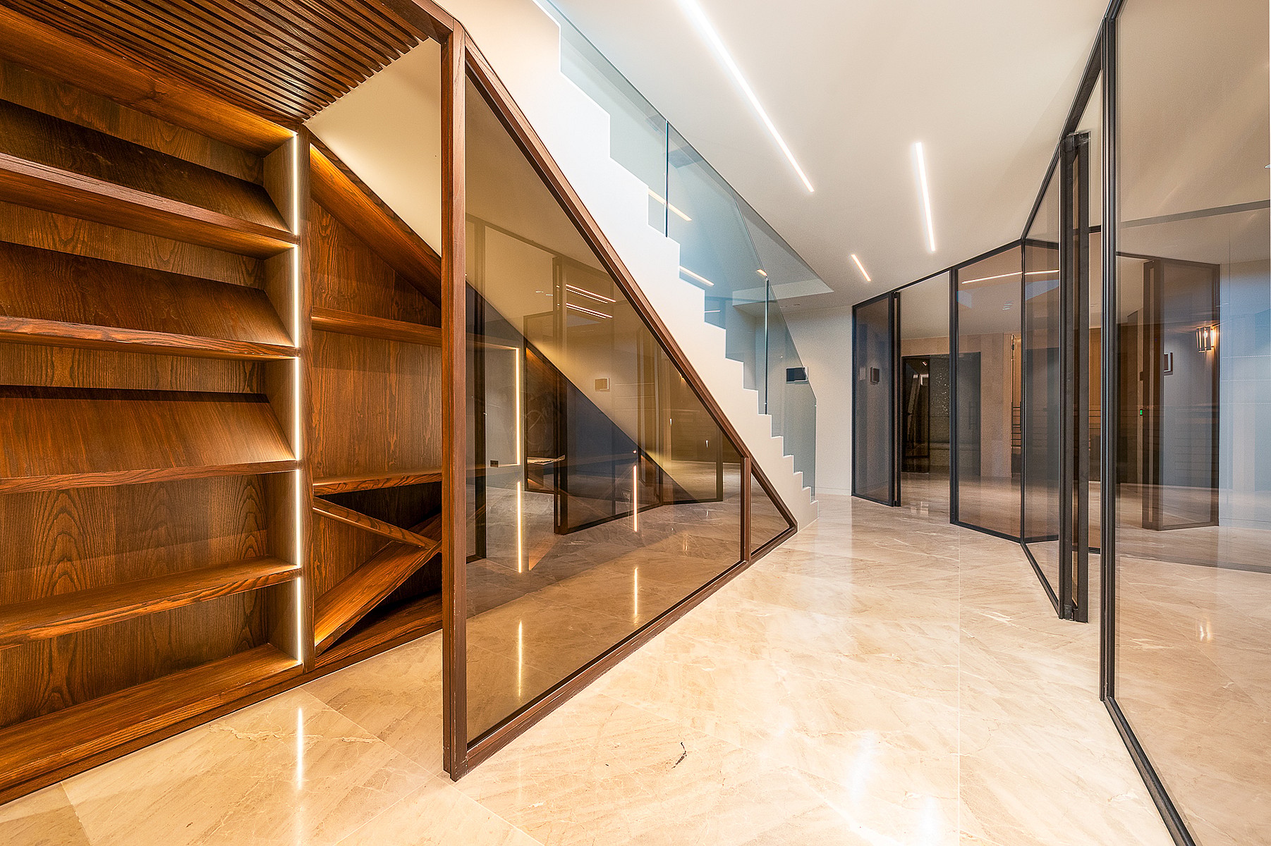 Interior hallway featuring polished marble floors, a glass staircase partition, and integrated wooden shelving units with linear lighting.