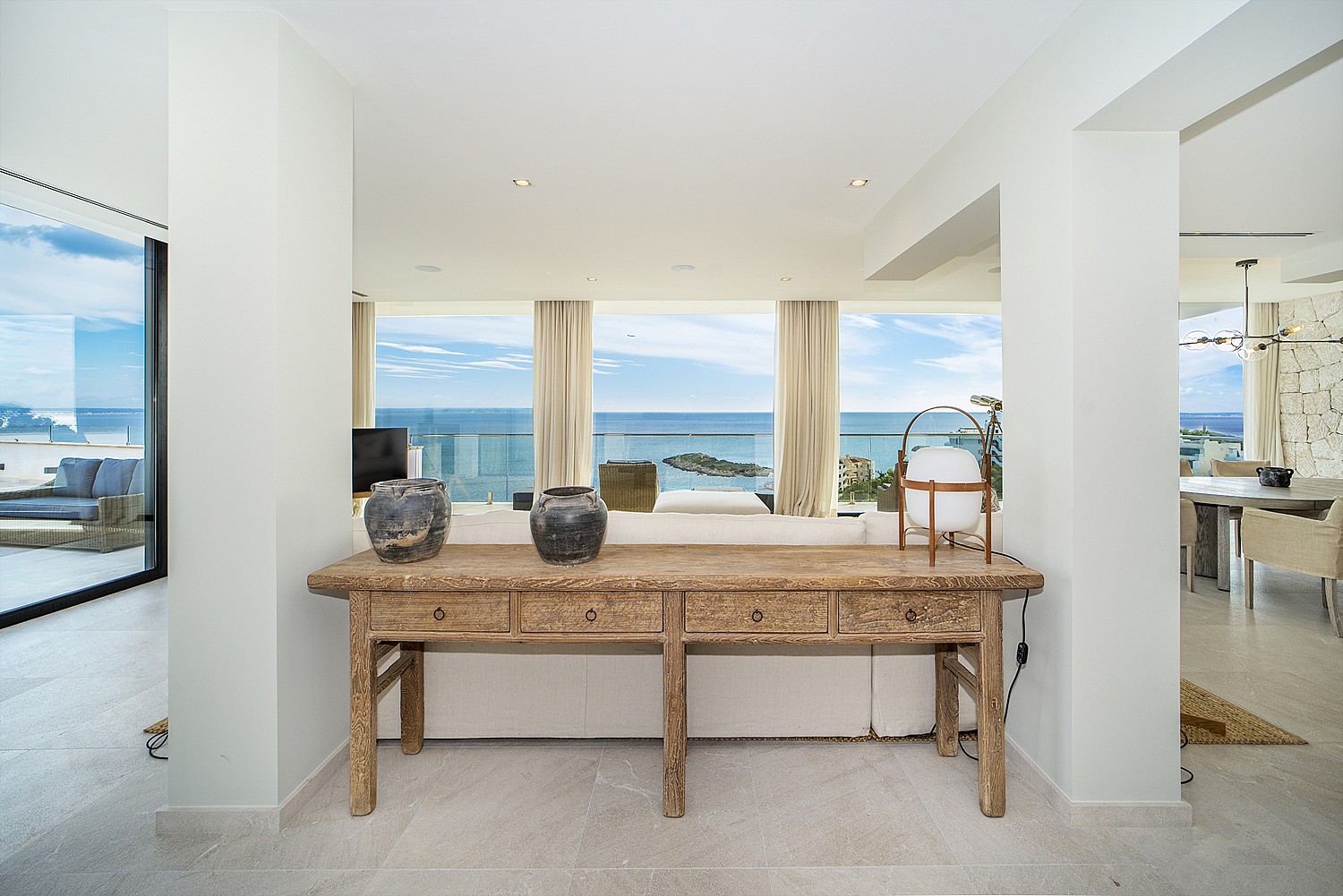 Interior view of an open-plan apartment featuring a rustic wooden console table, white walls, and floor-to-ceiling glass doors opening to a sea-facing terrace.