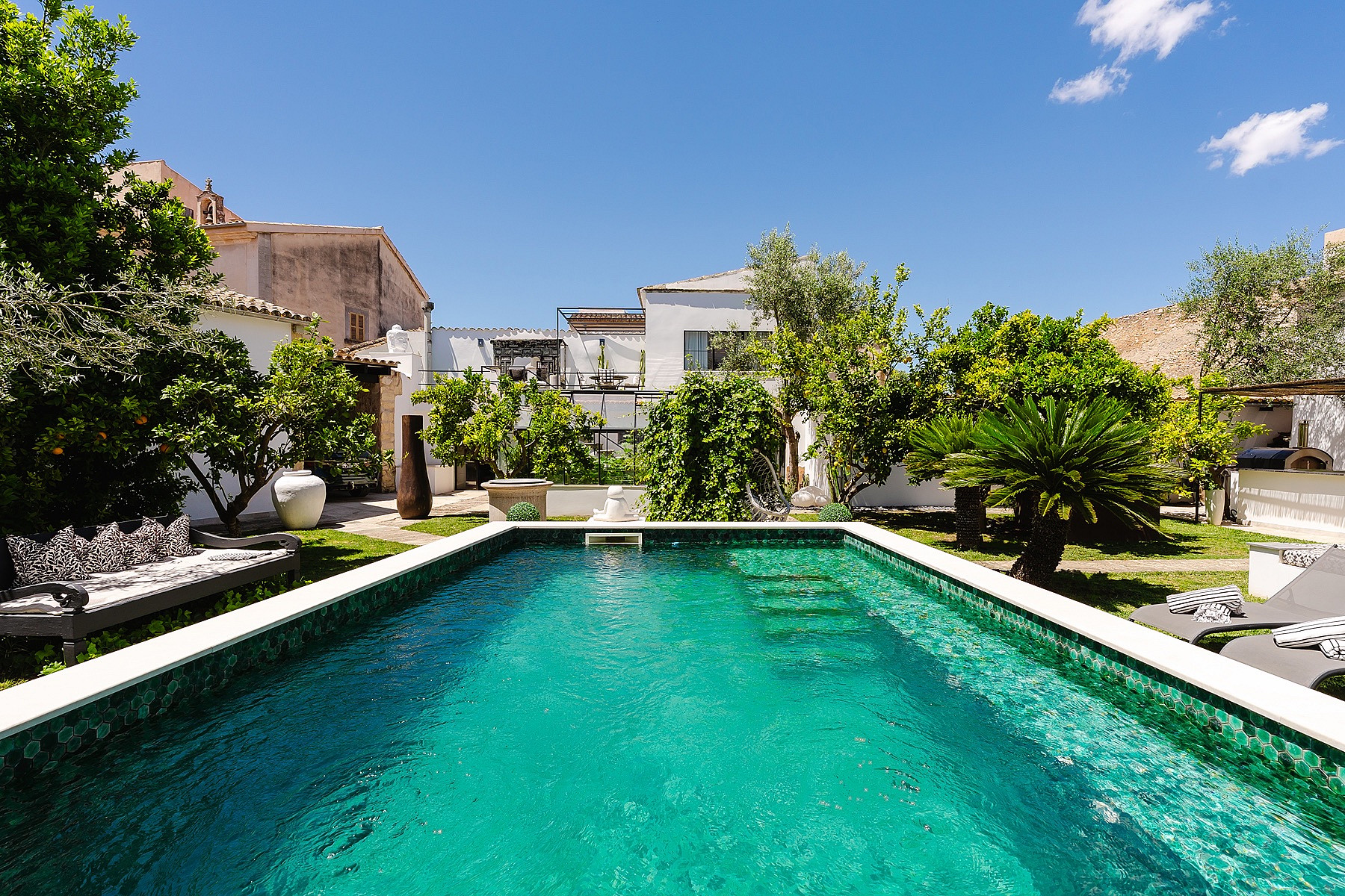 A rectangular swimming pool with turquoise mosaic tiles set within a lush, walled garden in Sencelles, featuring mature trees.