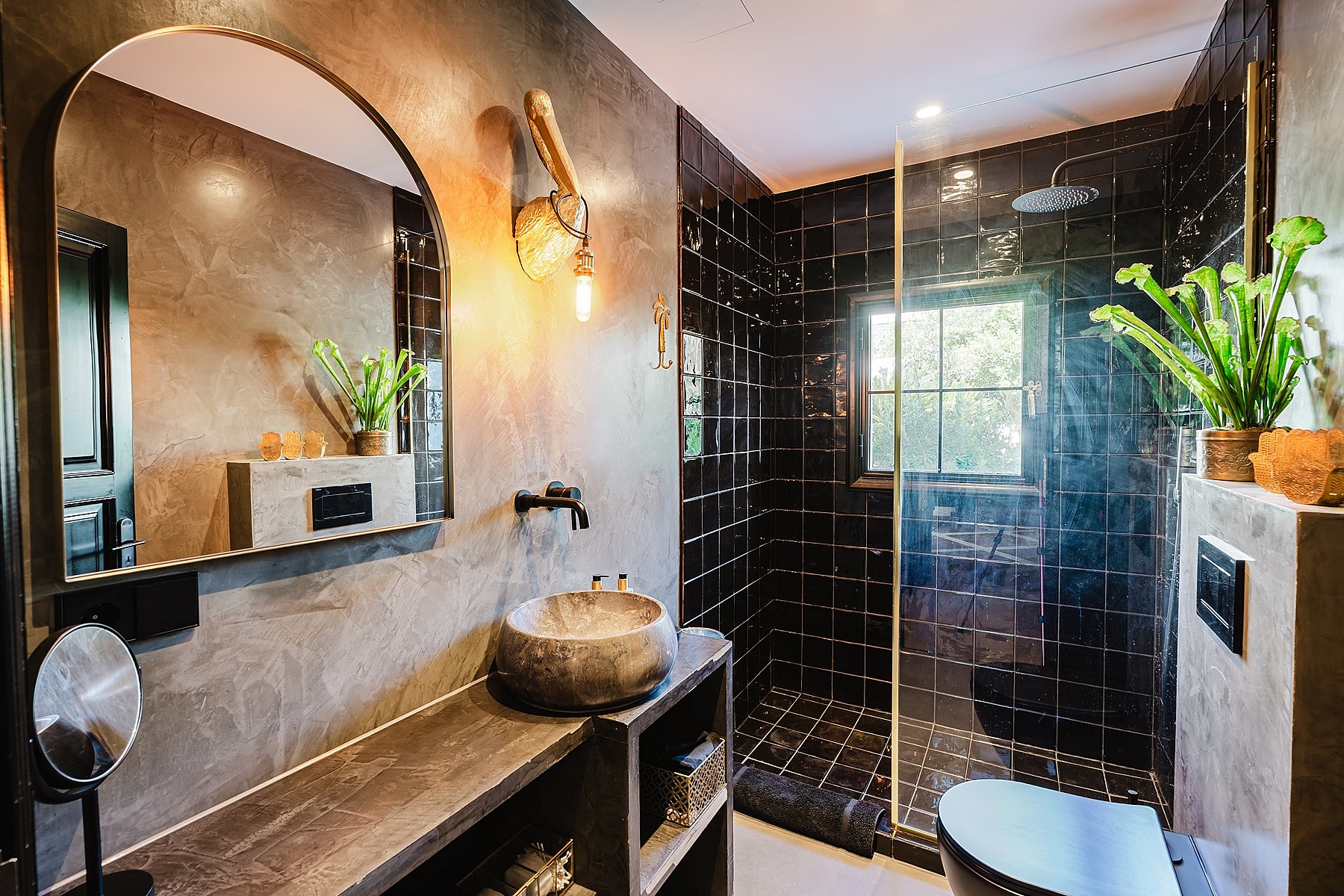 Contemporary bathroom with grey concrete walls, a dark stone washbasin, arched mirror, and a black tiled walk-in shower.