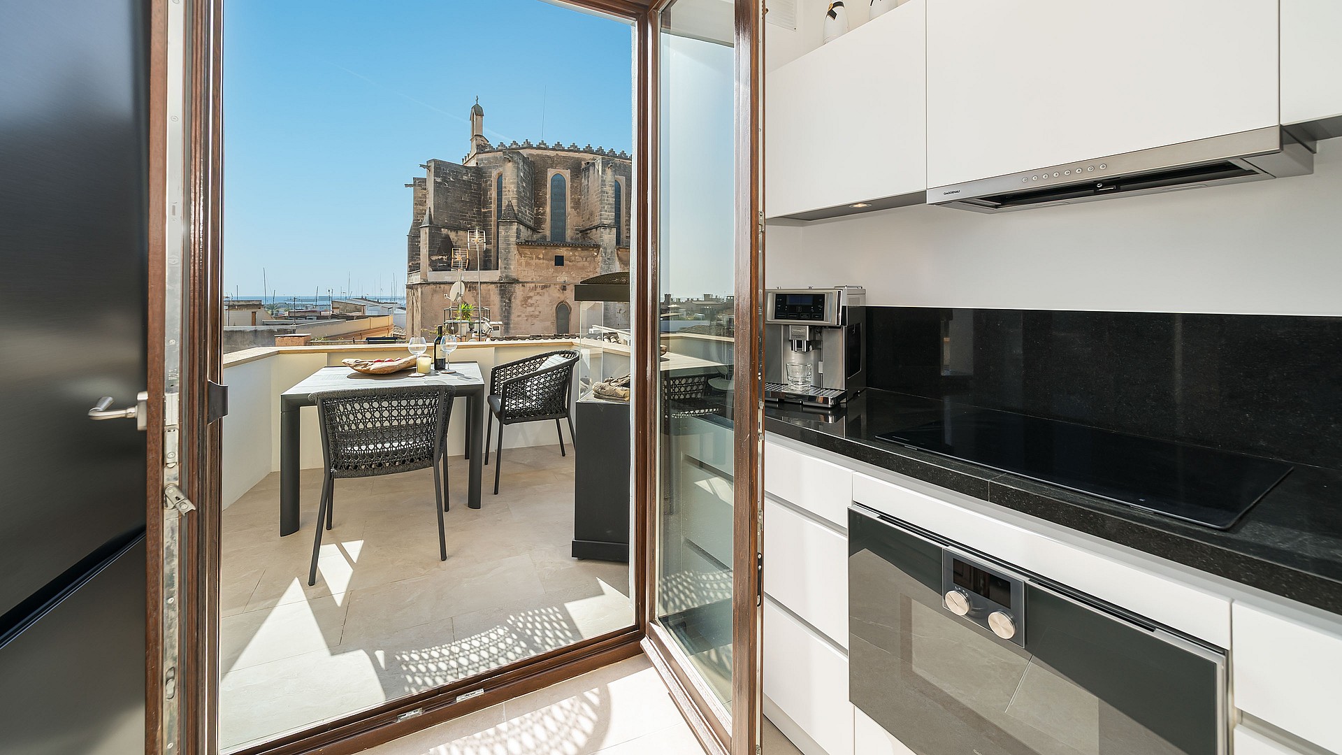 Modern white kitchen with black countertops opening to a sunny terrace with historic church views in Palma Old Town.